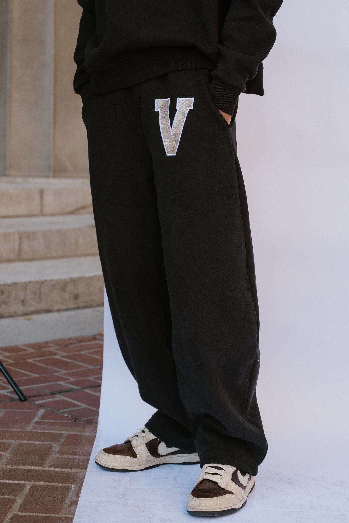 mens, A person wearing black wide-leg sweatpants with a large white outlined letter V on the left thigh, paired with brown and beige sneakers, standing outdoors against a white backdrop.