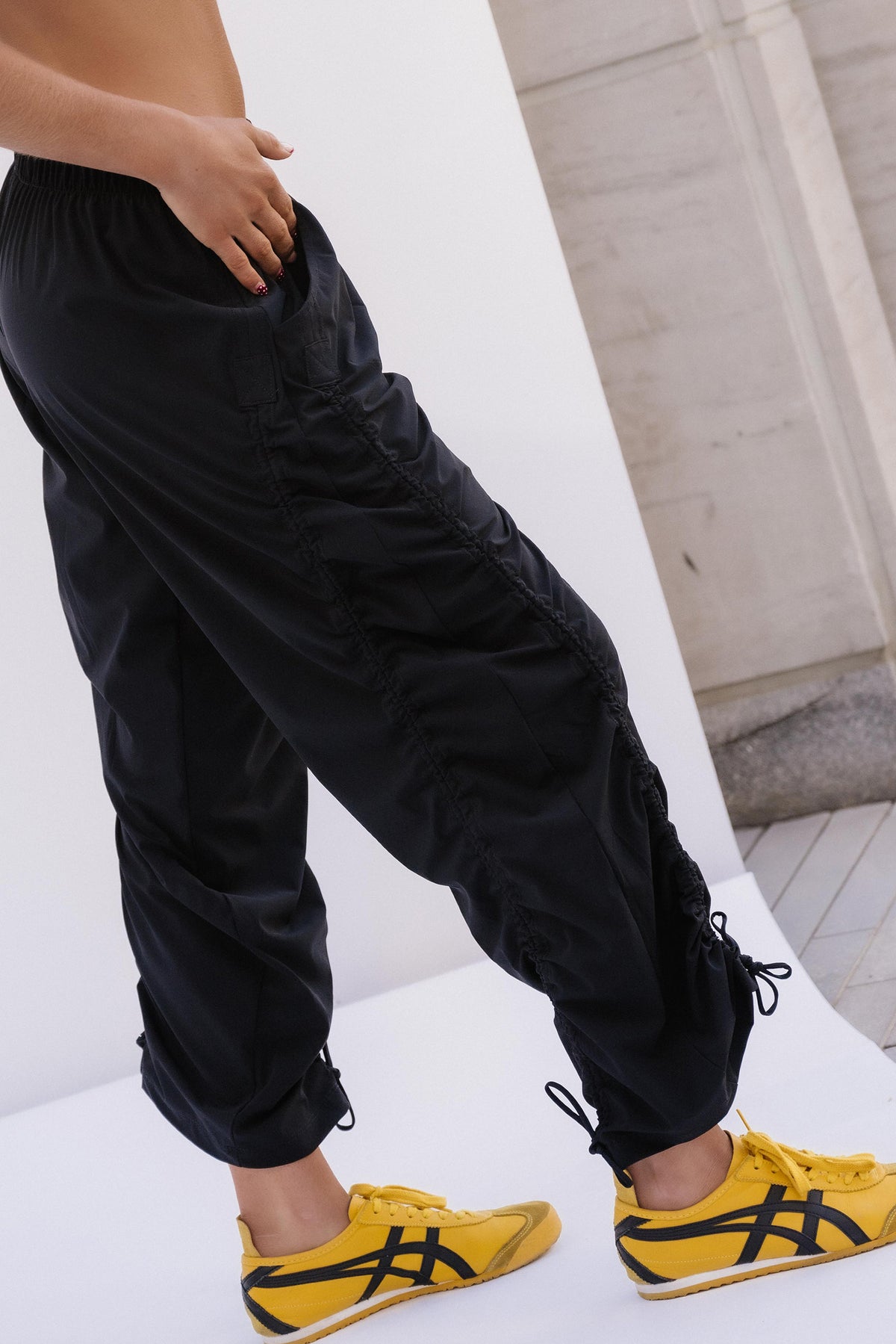 teen, A person wearing loose black drawstring pants and bright yellow sneakers with black stripes, photographed in a minimalist white and gray setting.
