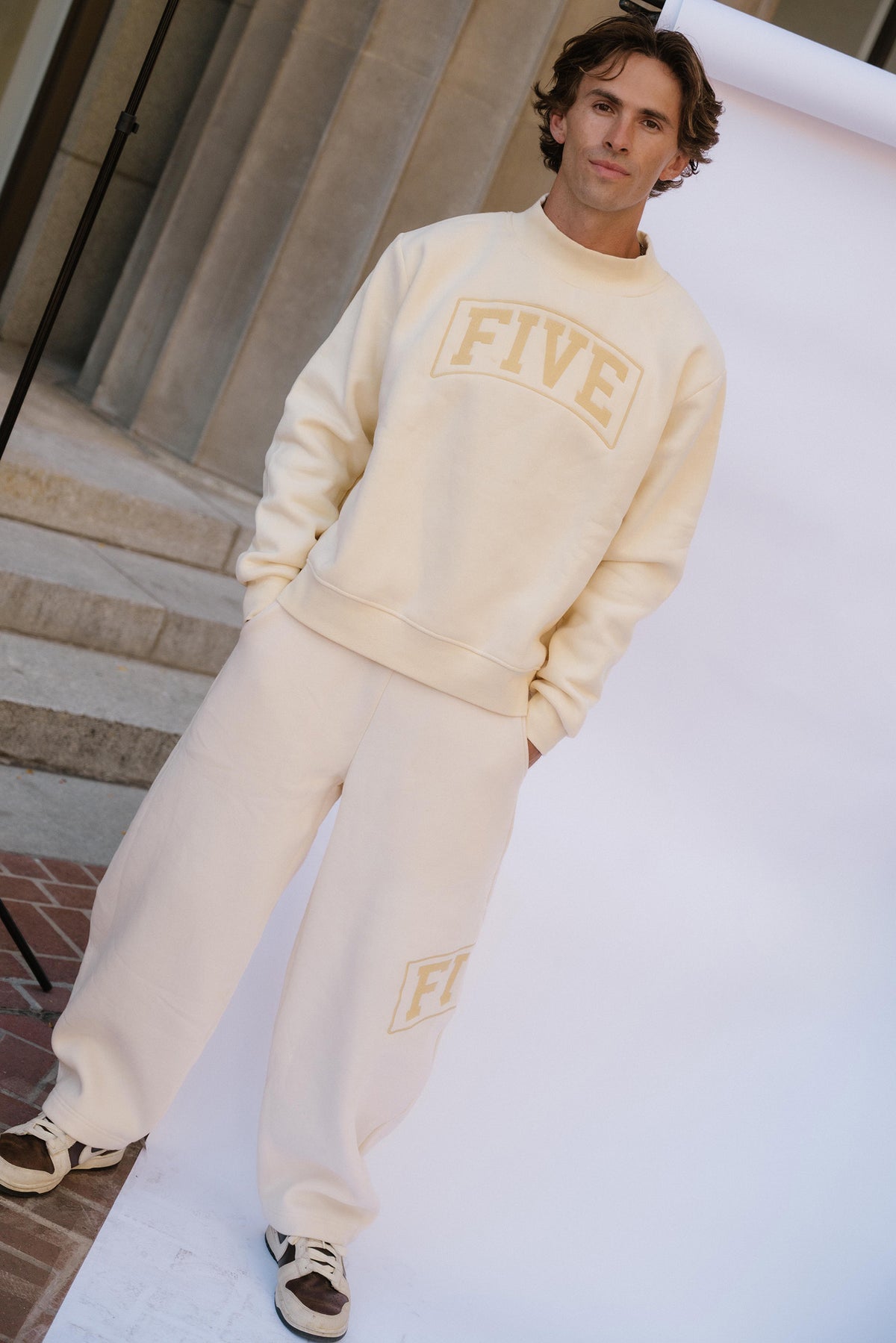 mens, A man modeling a cream-colored sweatshirt and sweatpants set with gold lettering that reads 'FIVE', standing in front of a white studio backdrop.