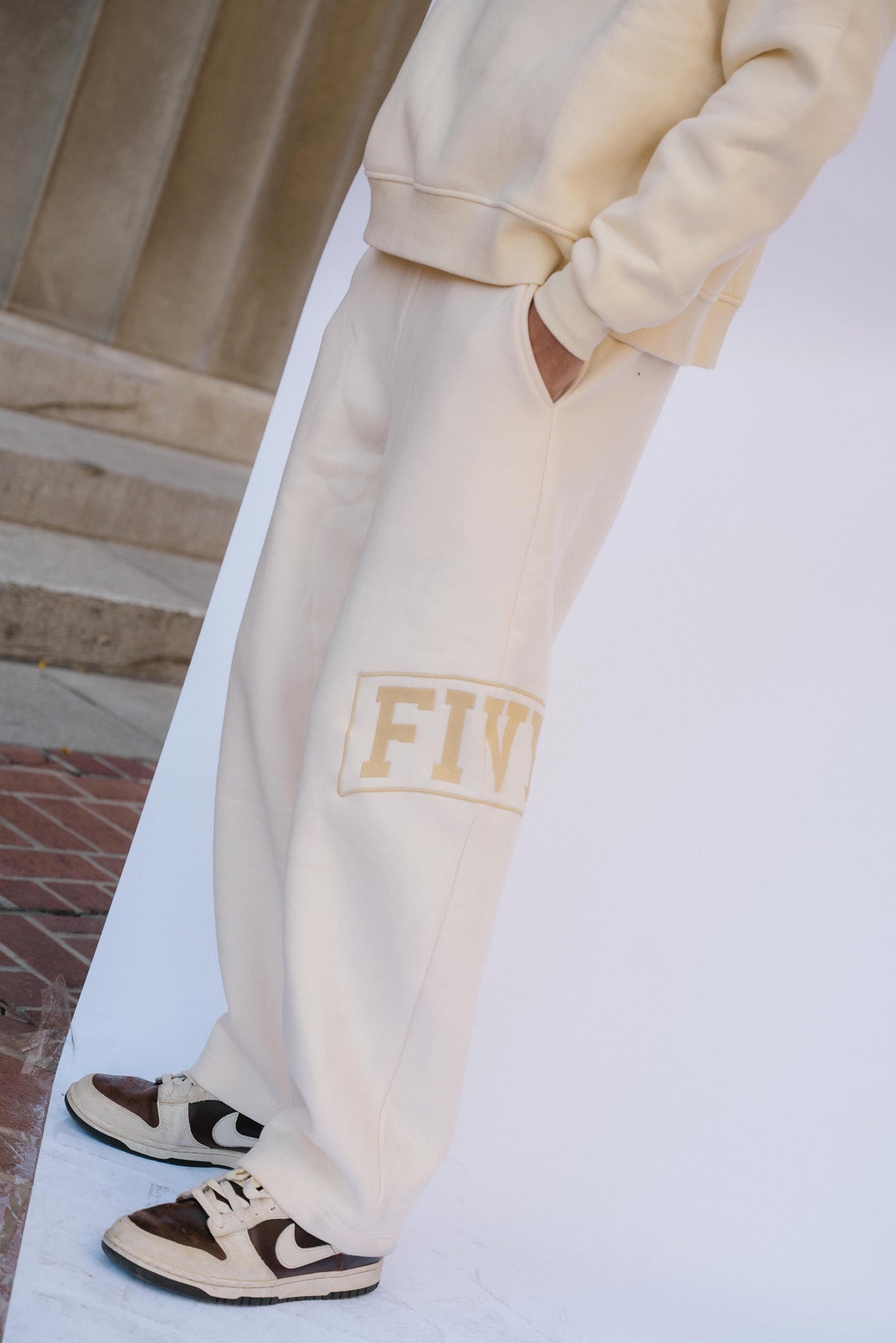 mens, A person wearing cream-colored sweatpants with a gold 'FIV' patch on the leg, paired with brown and cream sneakers, standing against a white backdrop.