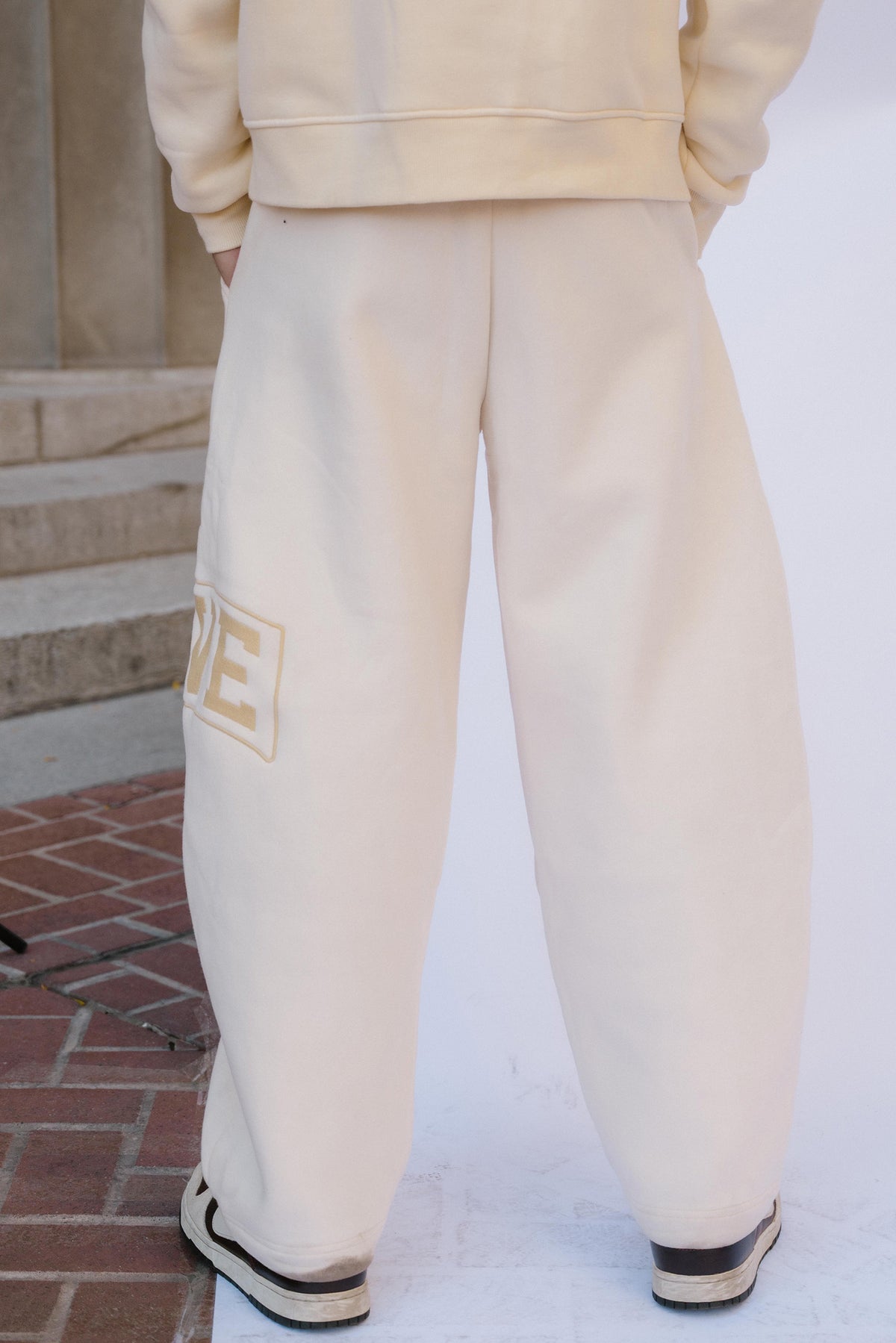 mens, A person shown from the back wearing cream-colored sweatpants with a gold embroidered patch on the left leg, paired with a matching cream sweatshirt and black-and-white sneakers, standing outdoors on a brick and concrete surface.