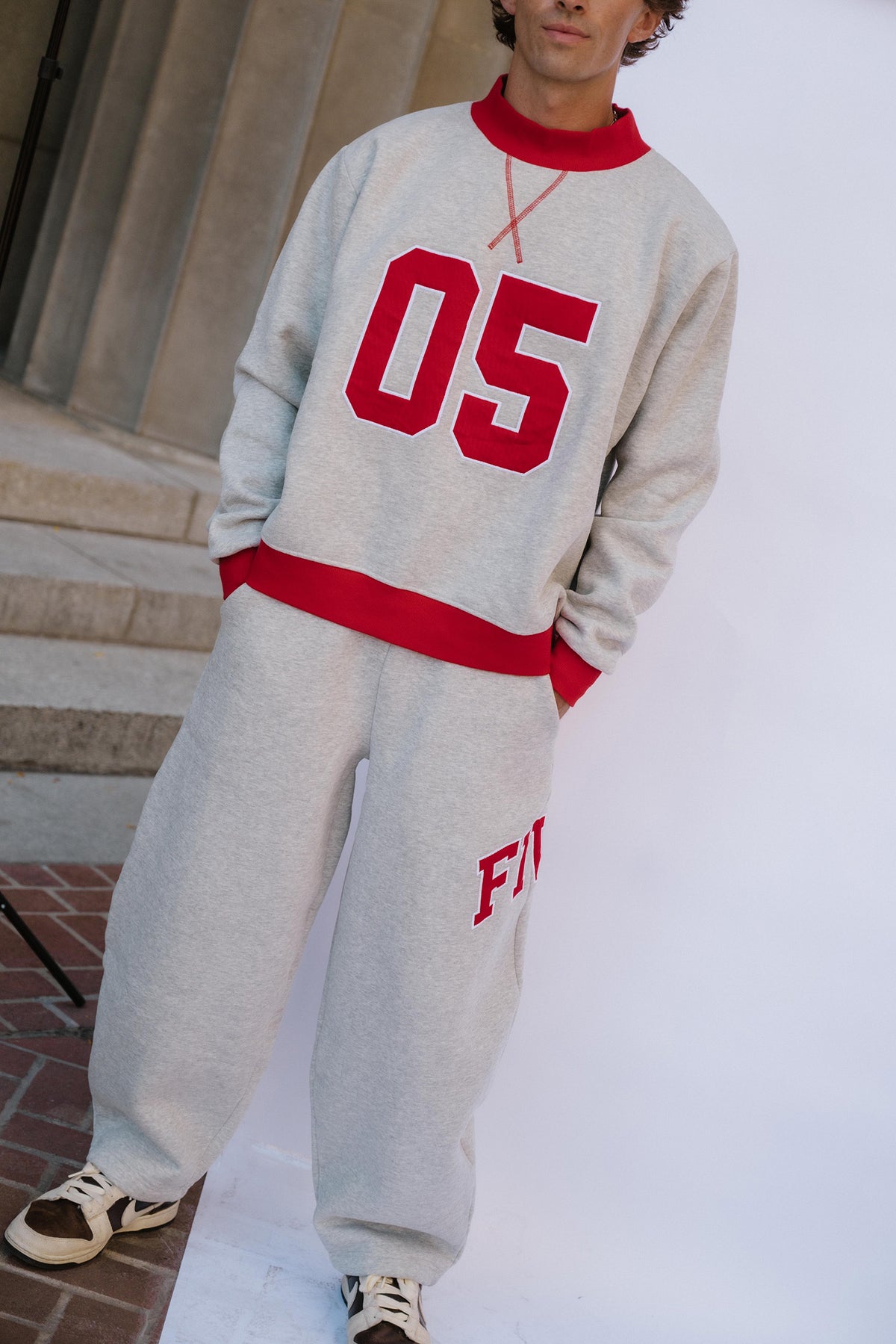 mens, A person wearing a light gray heather sweatshirt and matching sweatpants with bold red ribbed trim and red lettering, standing outdoors near stone steps and a white backdrop.