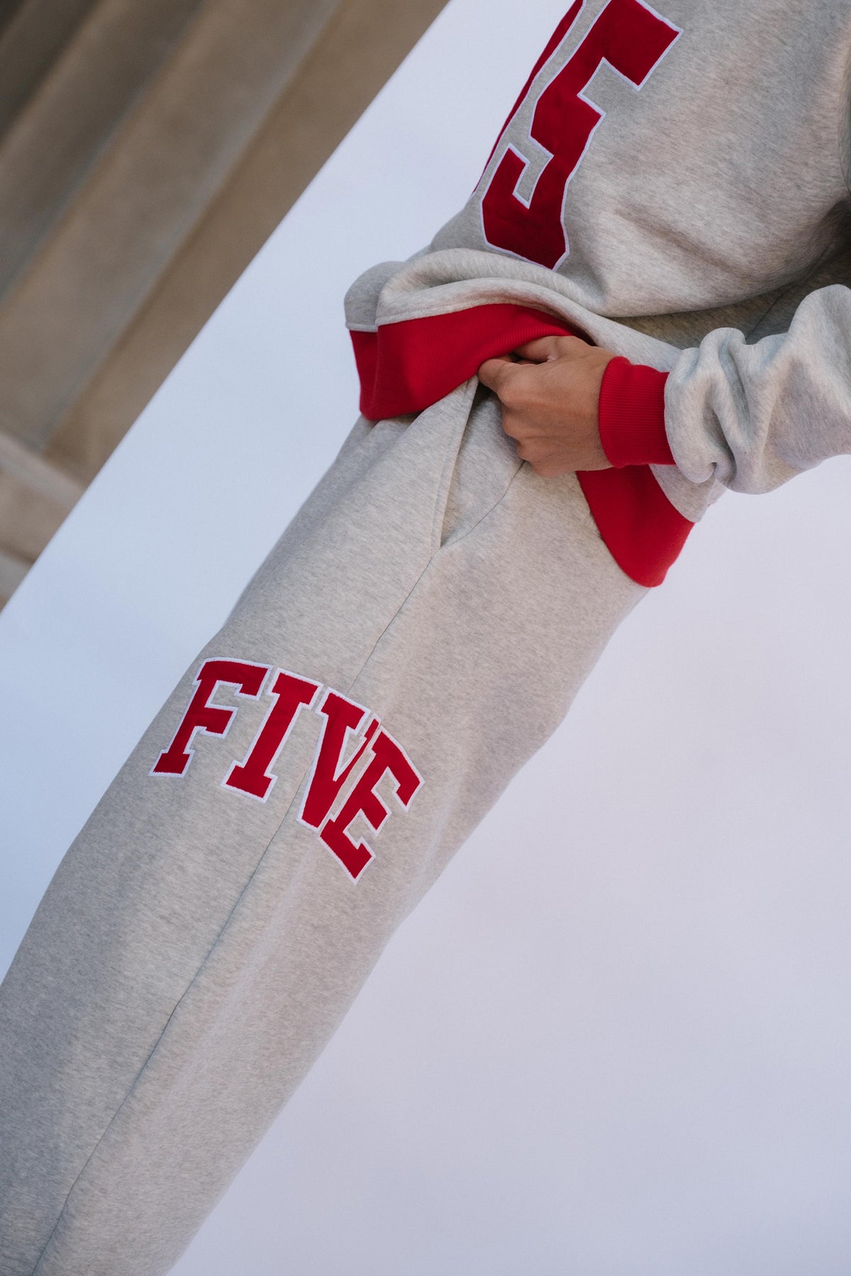 mens, Close-up of a person wearing light heather gray sweatpants with red trim and the word "FIVE" in red varsity-style lettering on the leg, holding up a matching heather gray sweatshirt with red trim and a large red number.