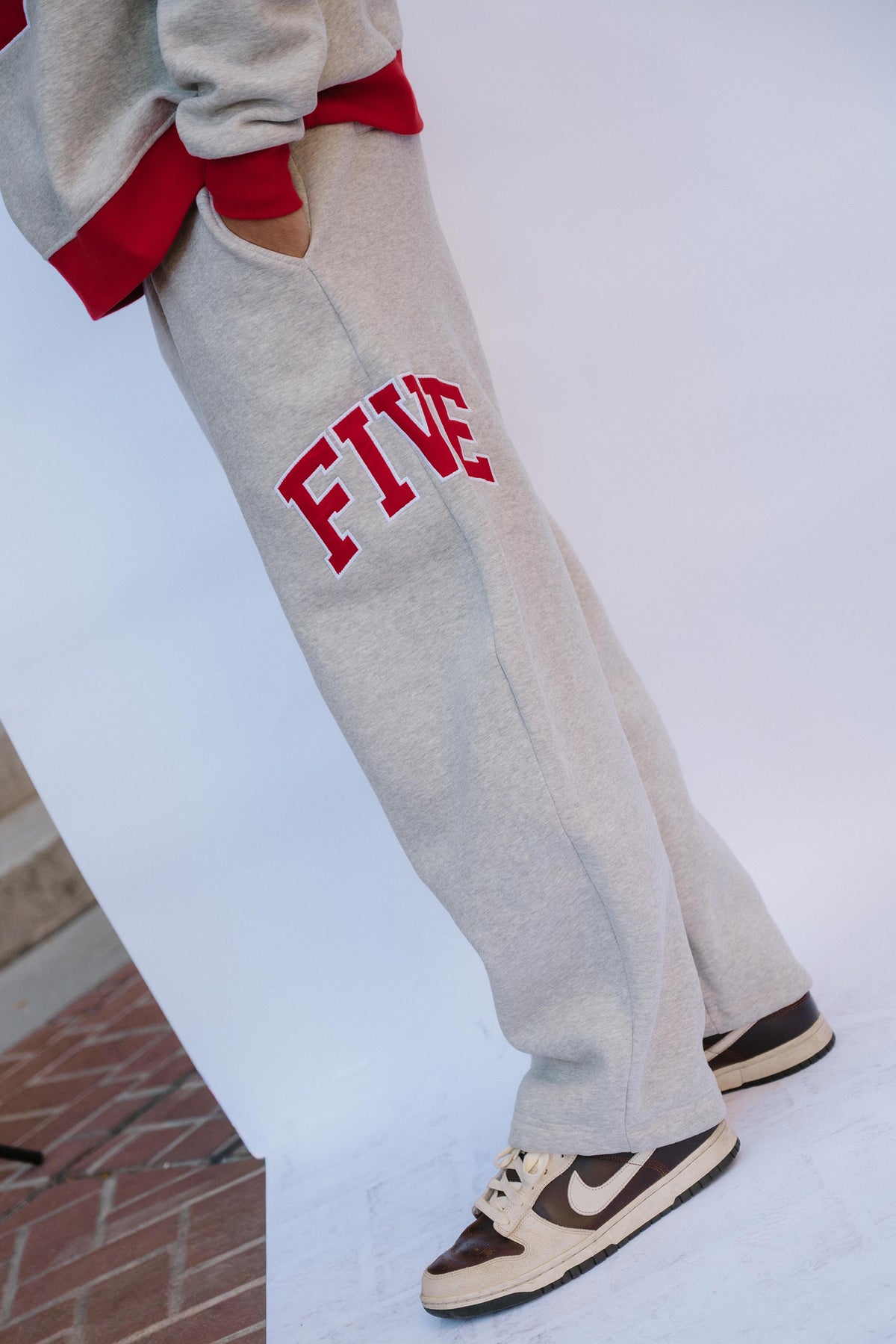mens, A person wearing light heather gray sweatpants with red collegiate-style trim and the word 'FIVE' in red on the leg, paired with brown and cream sneakers against a white studio backdrop.