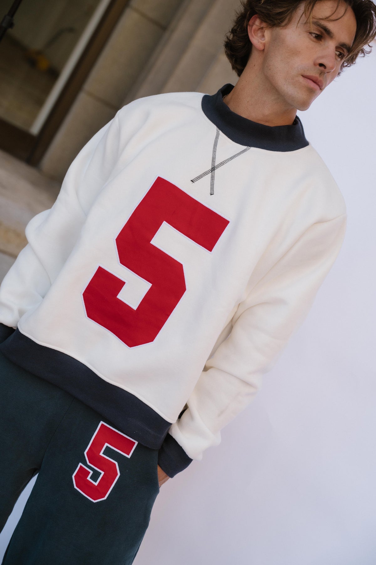 mens, A man wearing a cream-colored long-sleeve sweatshirt with a large red number 5 on the front, paired with dark navy pants featuring a matching red and white number 5 on the thigh.
