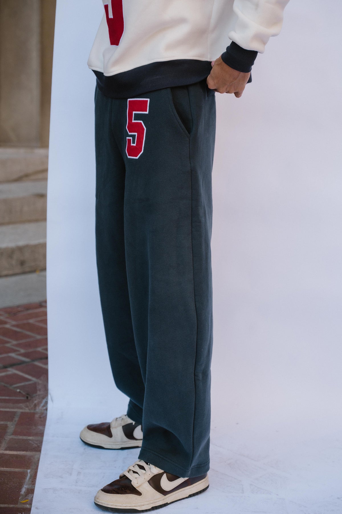 mens, A person wearing dark charcoal gray wide-leg sweatpants with a red and white number 5 on the left thigh, paired with a white sweatshirt and brown and white sneakers.