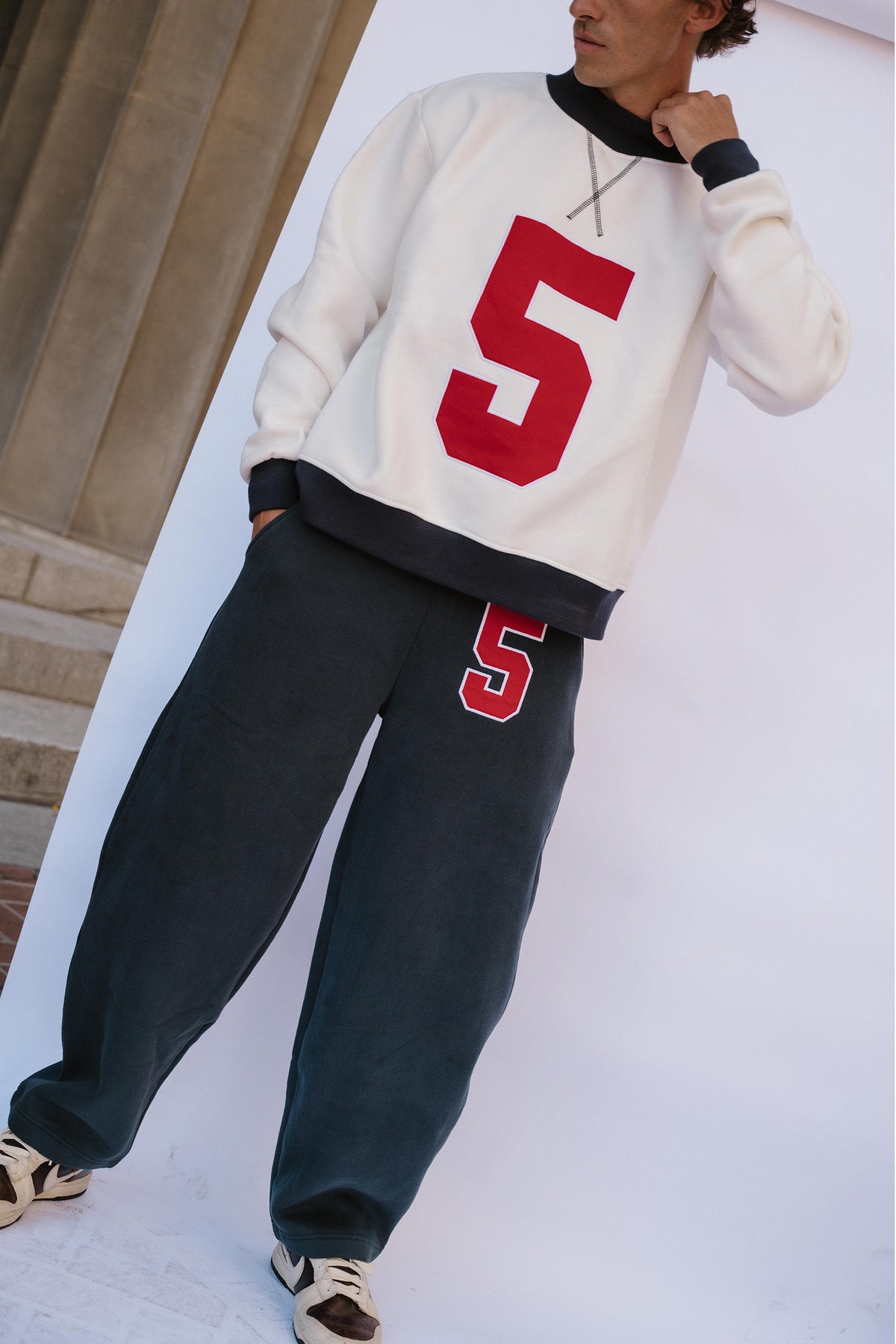 mens, A person wearing a cream sweatshirt with a large red number 5 on the front, dark charcoal sweatpants with a matching red and white number 5 on the thigh, and beige sneakers, posed against a white studio backdrop with a stone column visible on the left.