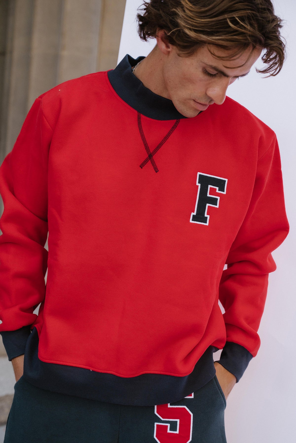 mens, A man wearing a bright red crewneck sweatshirt with a dark navy collar, cuffs, and hem, featuring a large navy block letter 'F' outlined in white on the left chest. He is looking down, with his hands in his pockets, wearing dark navy sweatpants.
