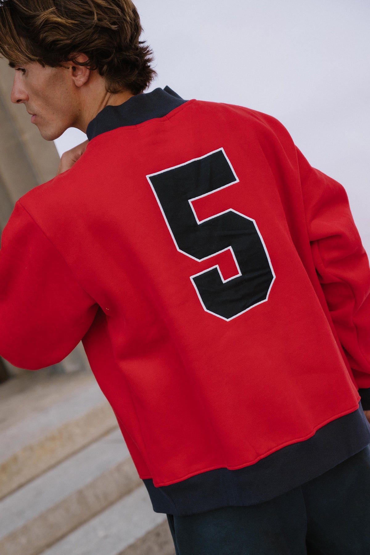 mens, A person wearing a bright red varsity-style jacket with a large black number 5 outlined in white on the back, photographed outdoors against a pale sky and stone steps.