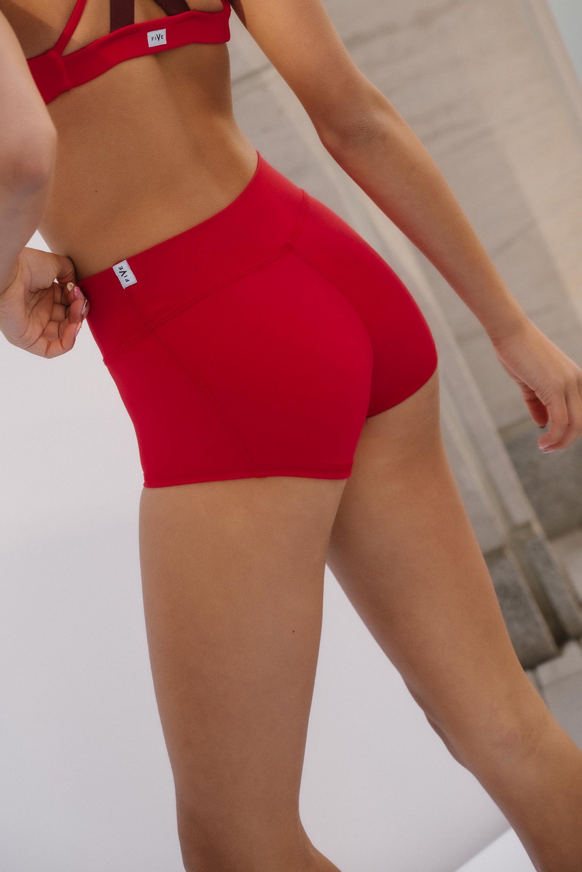 teen, close-up of a model wearing bright red athletic shorts and a matching red sports bra, with a small white brand tag visible on the waistband. 
