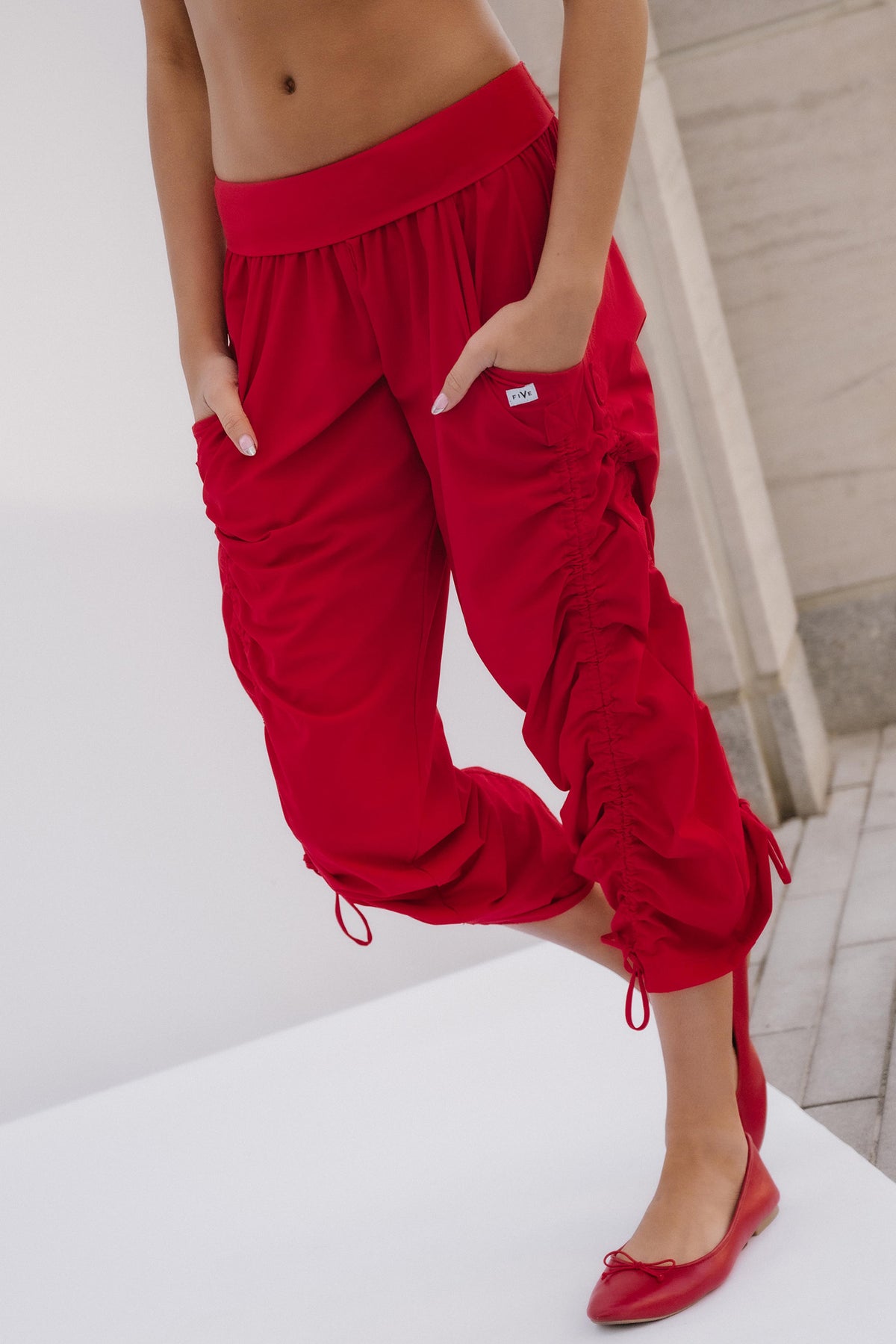 teen, A model wearing bright red ruched cargo-style pants with drawstring cuffs and side pockets, paired with matching red flats, shown against a clean white and light gray architectural background.