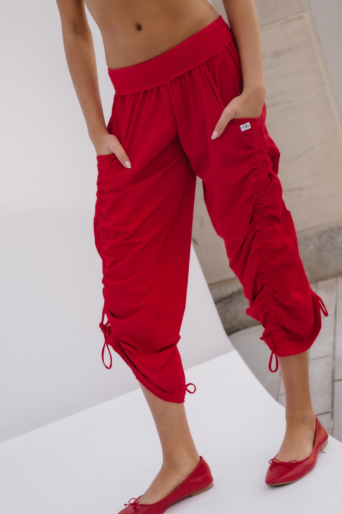 teen, A model wearing bright red ruched capri pants with side drawstrings and red ballet flats, shown from the waist down in a studio setting.