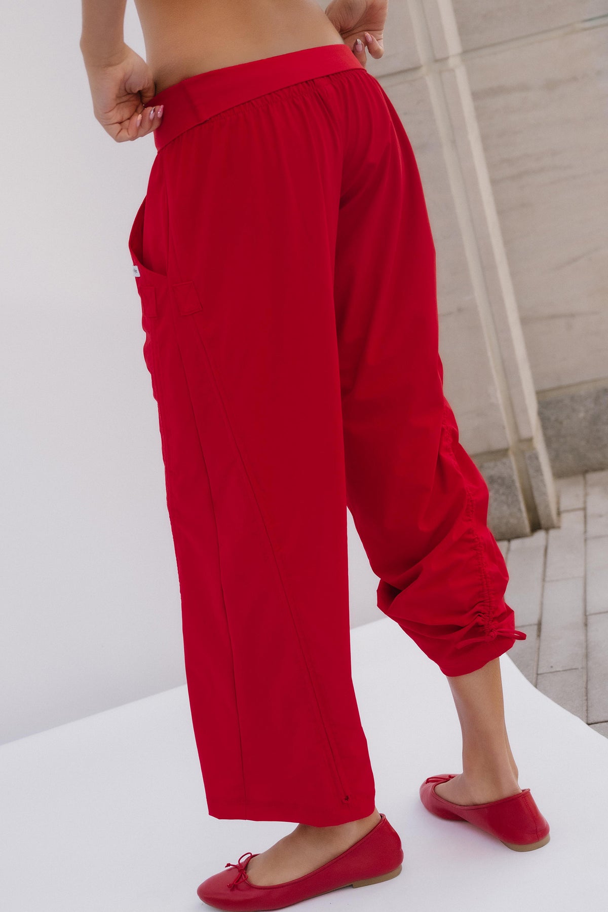 teen, A person wearing bright red cropped pants with a gathered elastic waist and rolled cuffs, paired with matching red flats, photographed indoors against a light neutral wall and floor.