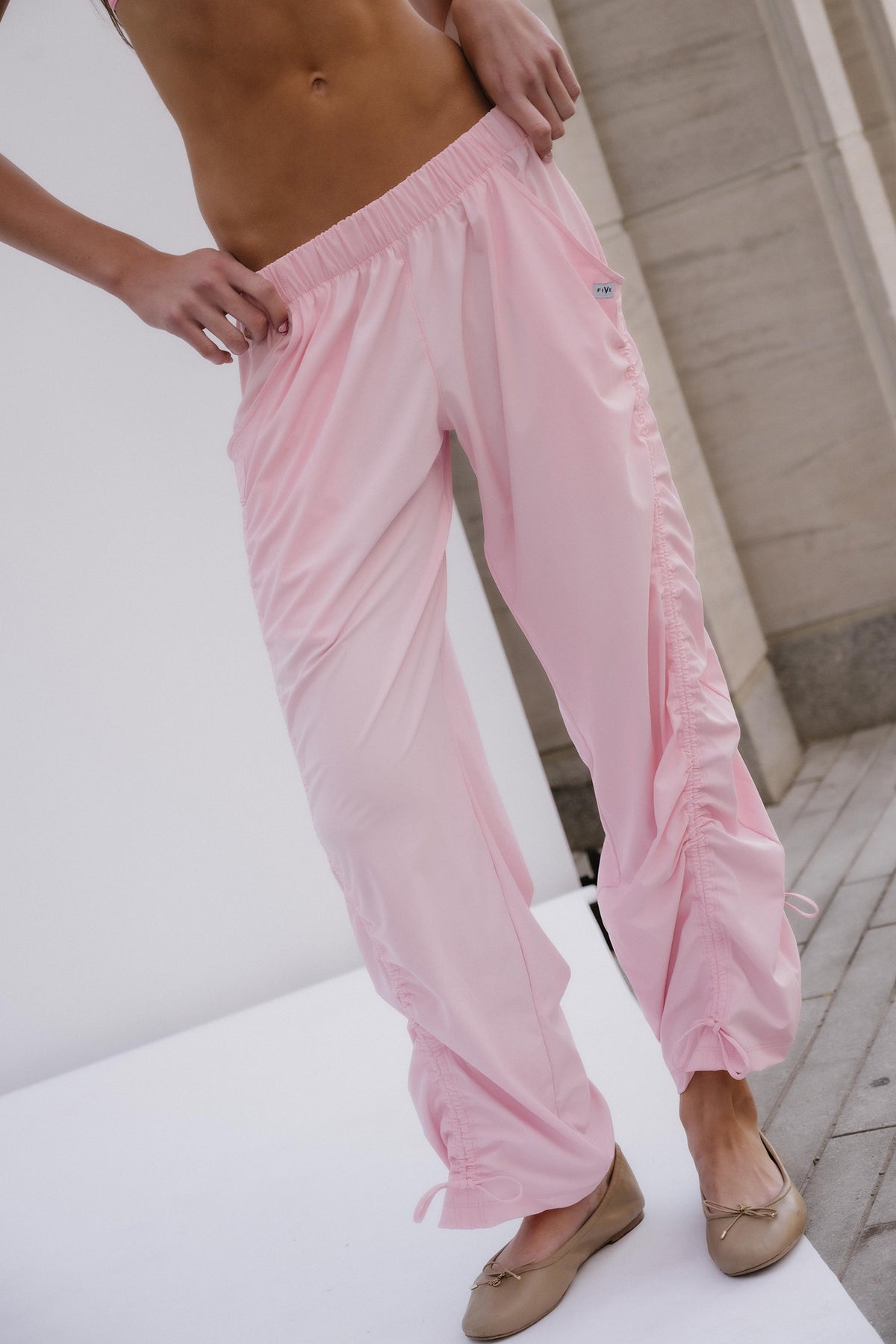 teen, A person wearing light pink wide-leg lounge pants with an elastic waistband and gathered cuffs, paired with beige flats, shown in an outdoor setting with a white wall and stone steps.