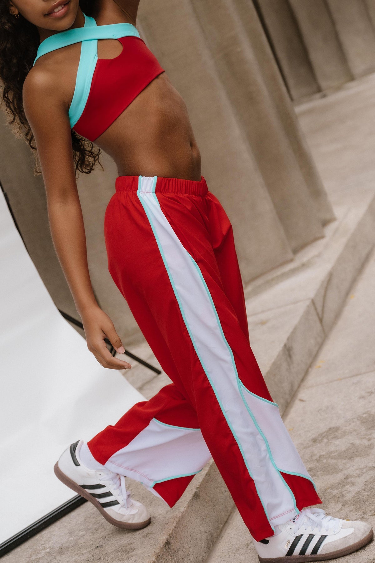 kids, kid wearing red and white athletic track pants with a light blue side stripe, paired with a matching red sports bra and white sneakers, posing on outdoor stone steps.