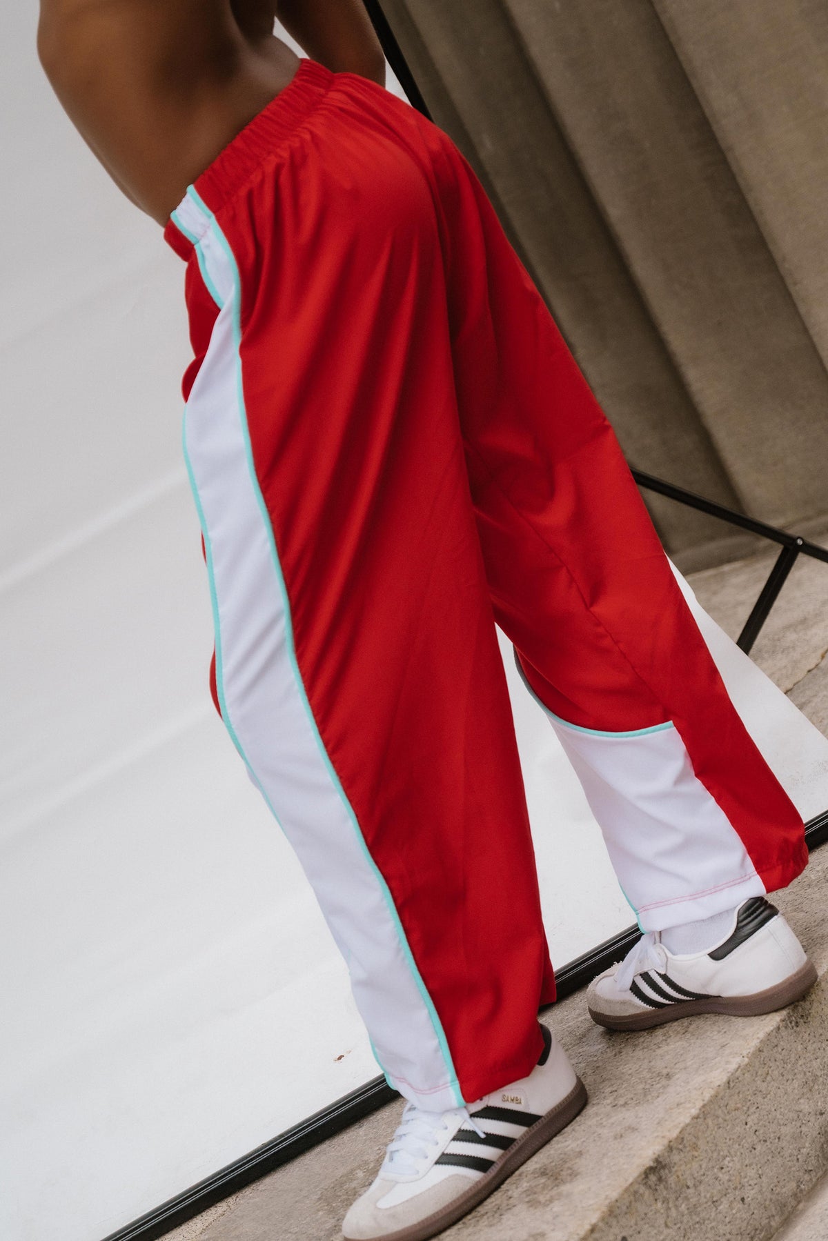 kids, A person wearing red track pants with white side panels and light blue piping, paired with white sneakers, photographed in a real studio-like setting with visible floor and backdrop.