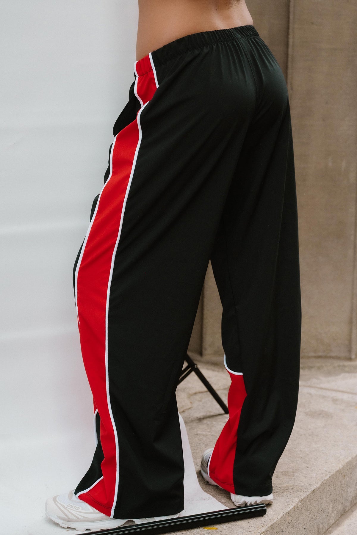 teen, Black wide-leg track pants with bold red and white side stripes, shown on a model from the waist down against a studio backdrop.