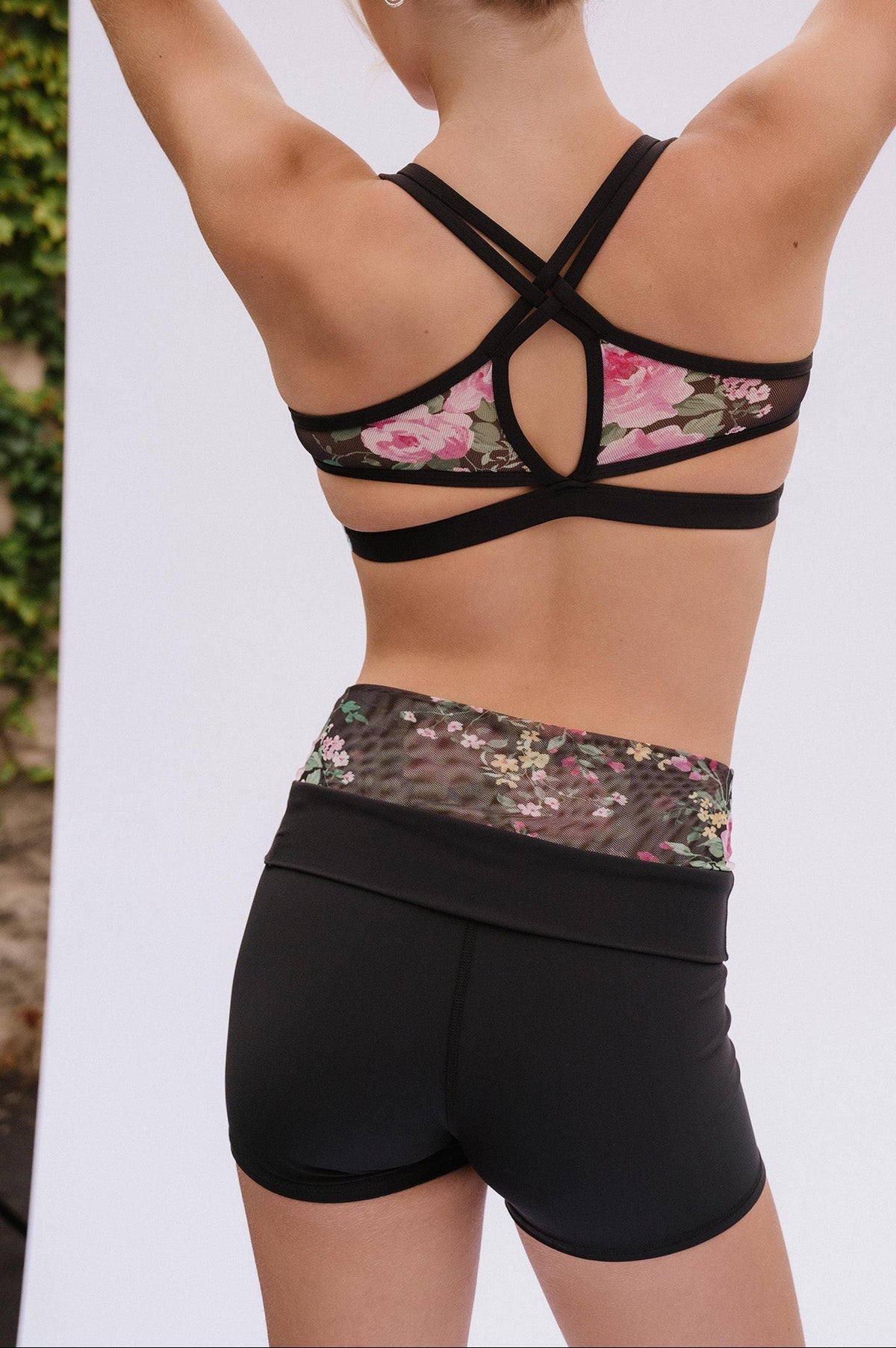 teen, shown from behind wearing a black sports bra and matching black athletic shorts with a floral mesh waistband, standing against a white studio backdrop.
