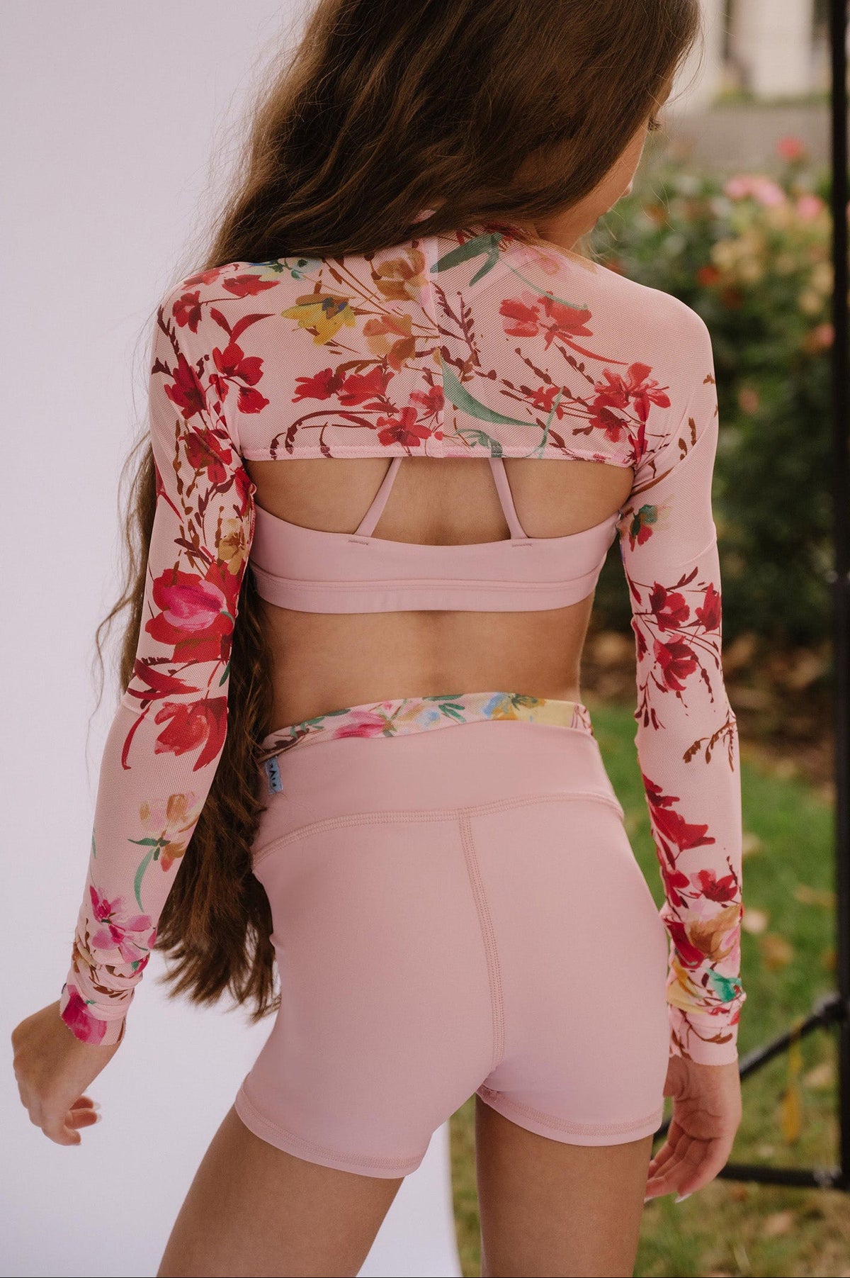 kids, A girl seen from behind wearing a pink floral long-sleeve crop top and matching pink athletic shorts, standing outdoors near a white studio backdrop and a black stand.