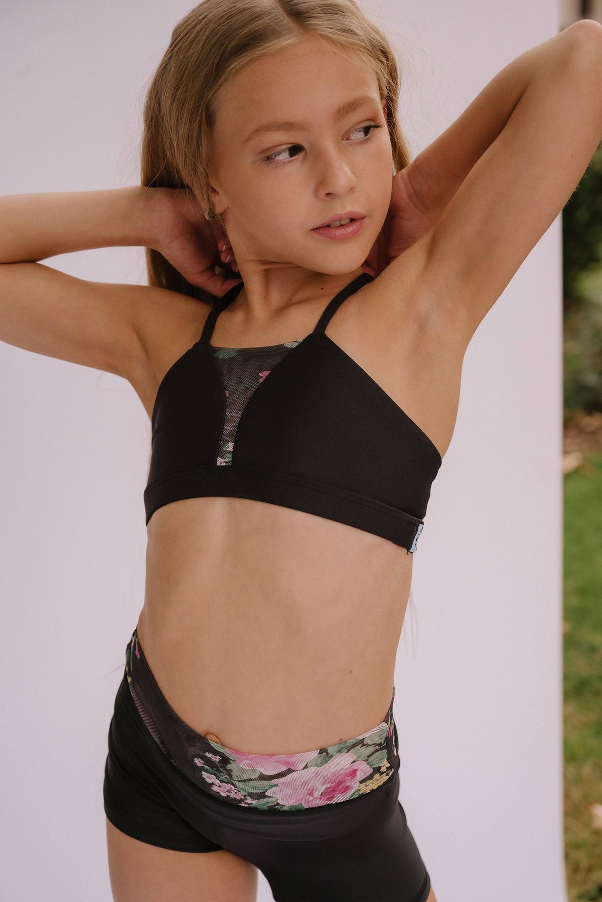 kids, A young girl modeling a black two-piece athletic set featuring a sports bra with sheer mesh side panels and matching high-waisted shorts with a pink and green floral waistband.