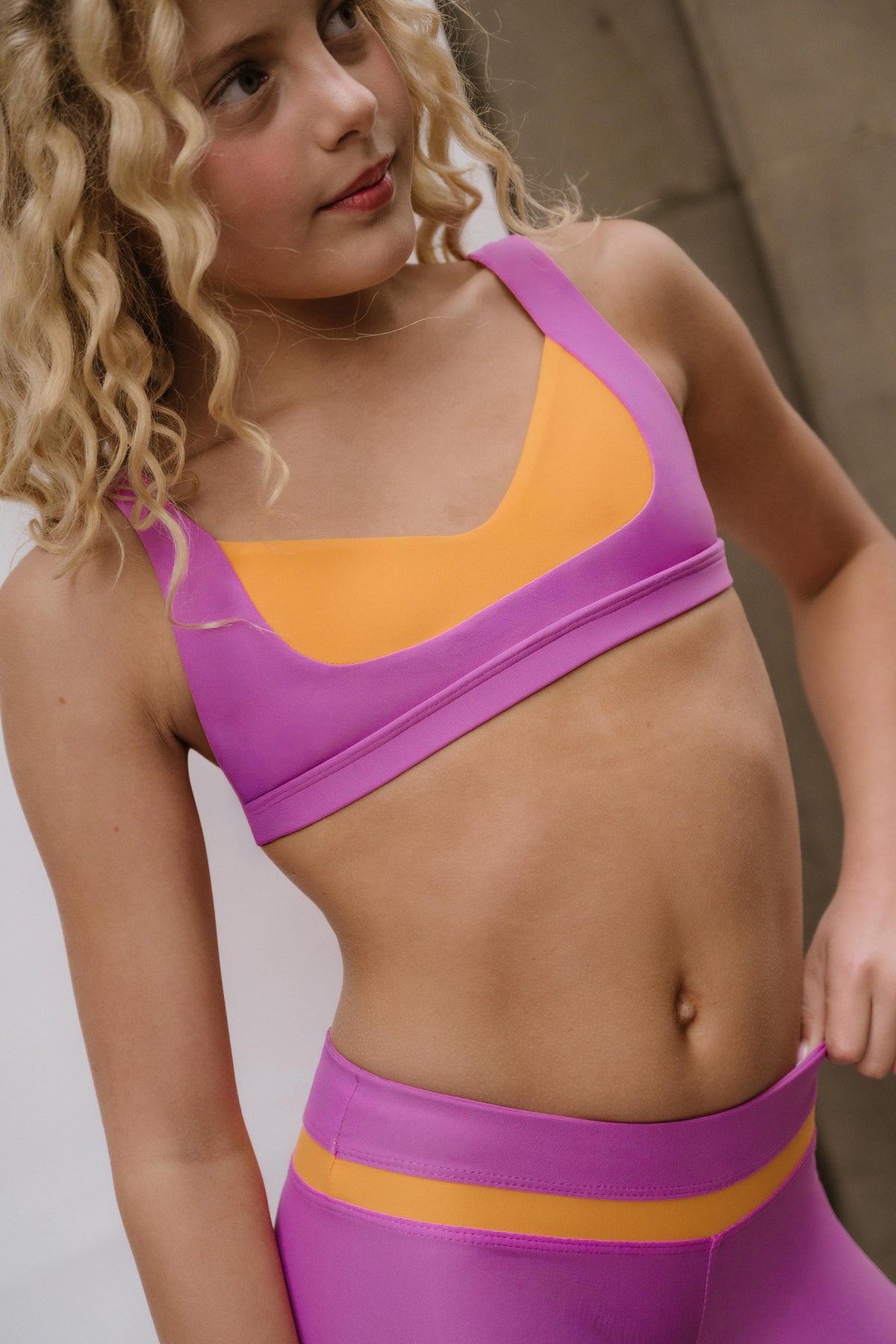 kids, kid A close-up of a young girl wearing a two-tone athletic sports bra and matching high-waisted leggings in purple and orange, with her blonde curly hair visible.