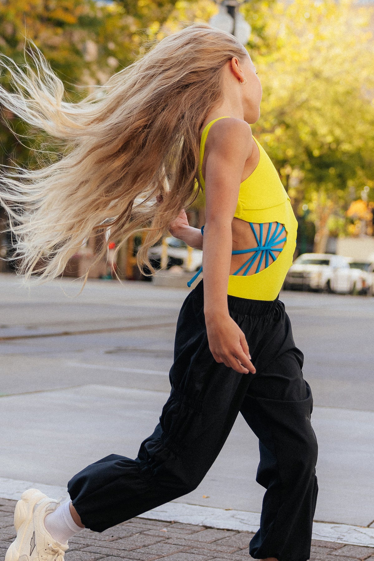 kids, A young girl with long blonde hair runs across a city street wearing a bright yellow leotard with a blue sunburst graphic on the side, paired with loose black pants and white sneakers.