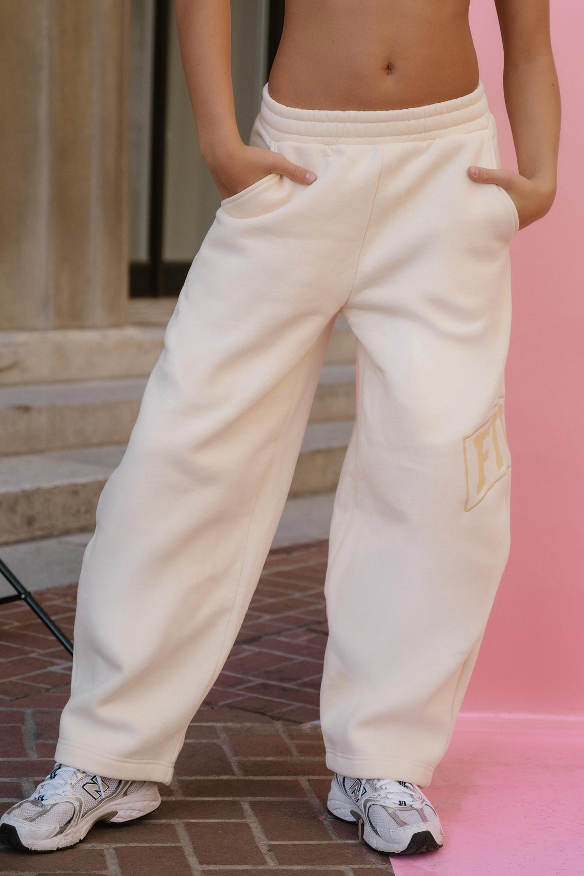 teen,  person wearing oversized cream sweatpants with side pockets and a gold embroidered 'F' patch on the lower left leg, paired with white sneakers, standing against a pink backdrop.