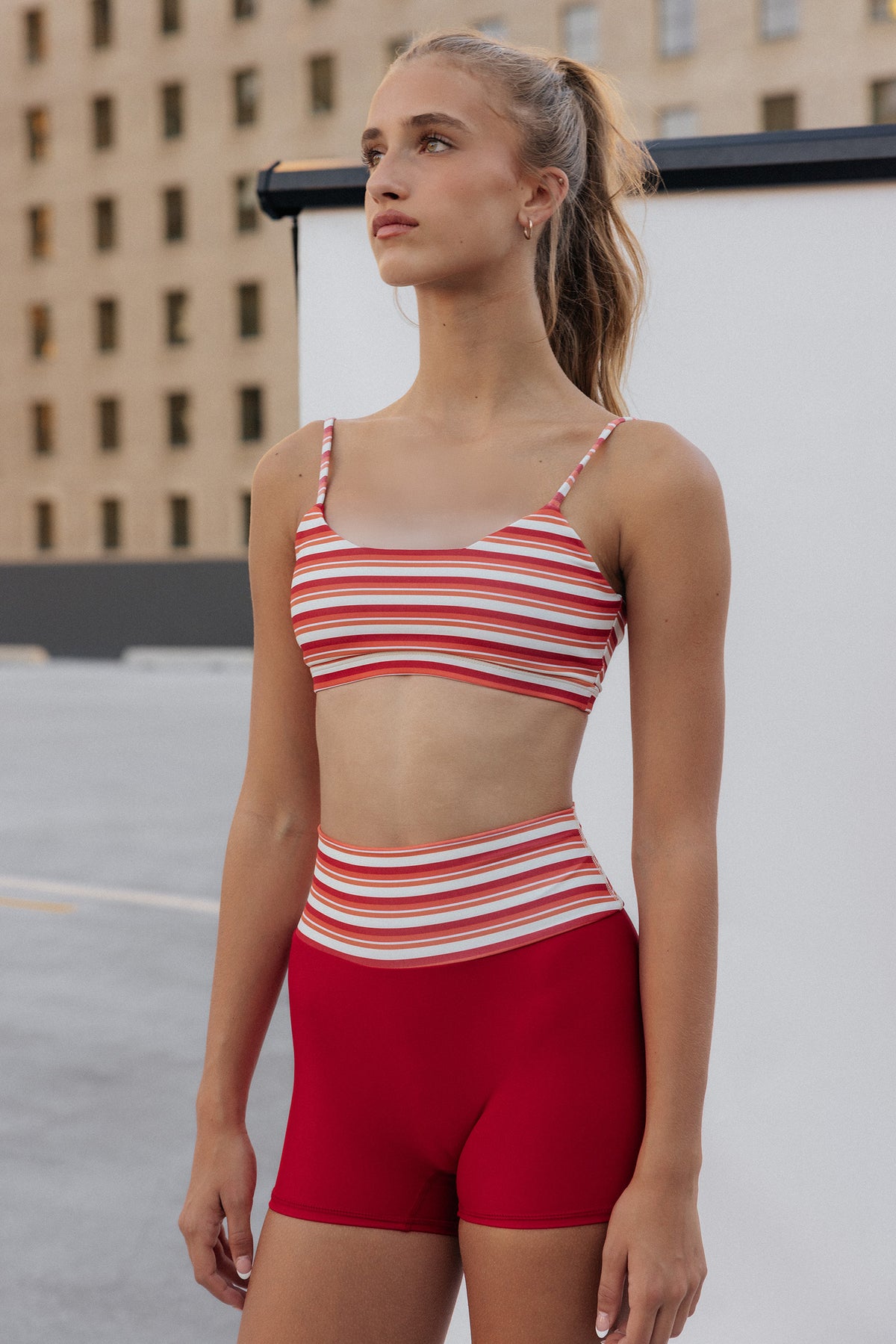 teen, girl modeling a matching two-piece athletic set with a red, white, and orange striped sports bra and solid red high-waisted shorts, set against an outdoor urban background.