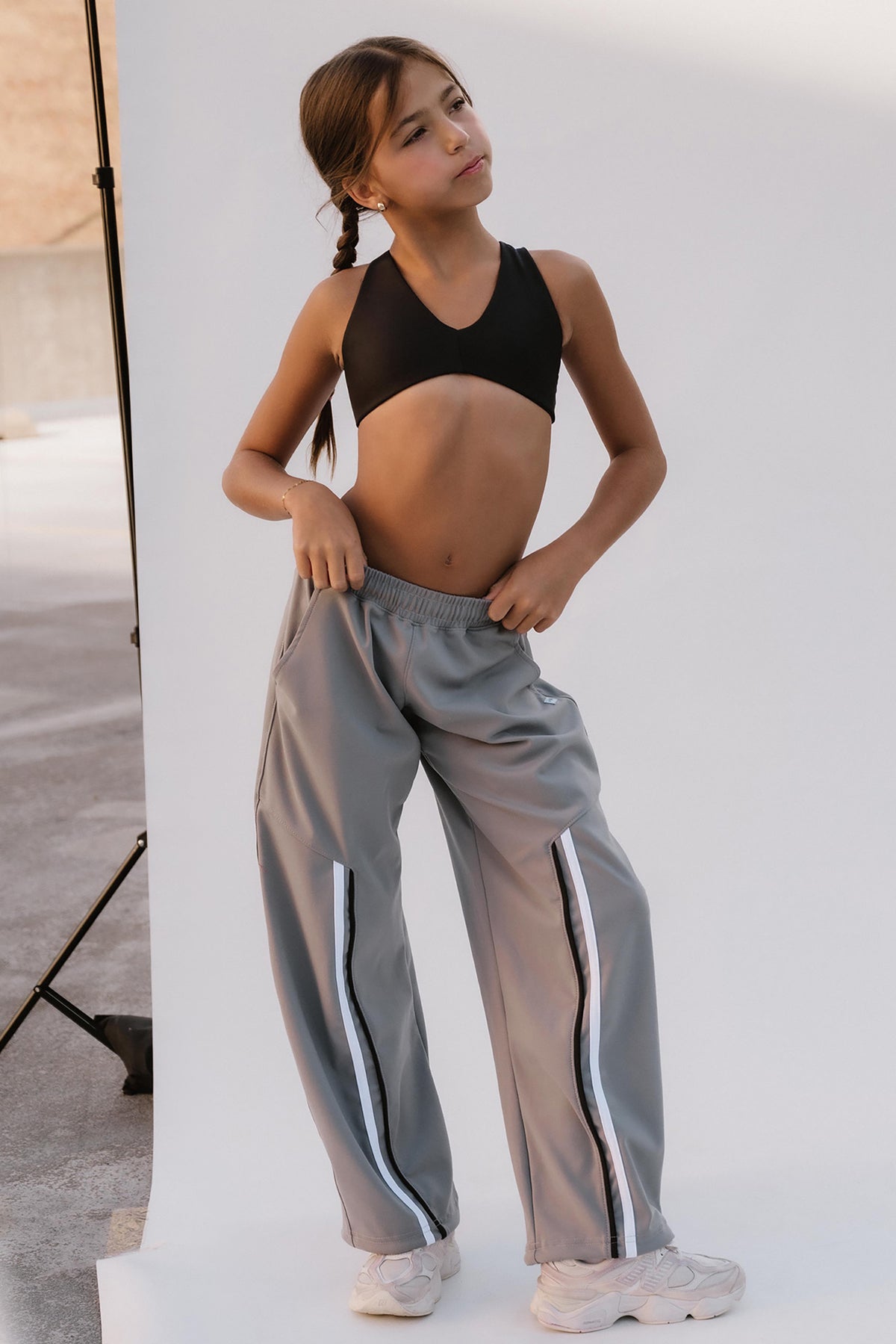 kids, A young girl modeling a pair of loose-fit gray sweatpants with white side stripes, paired with a black sports bra and white sneakers, standing against a white studio backdrop.