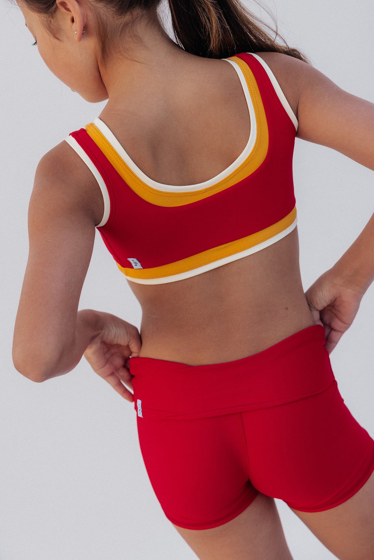 kids, A child wearing a red sports bra and matching red athletic shorts, shown from the back against a light gray studio background.