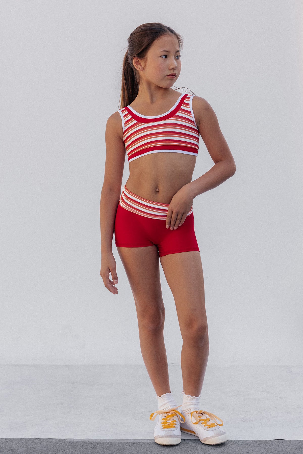 kids, A young girl stands in a studio wearing a red and white striped sports bra and matching red athletic shorts, paired with white socks and white sneakers with yellow laces.