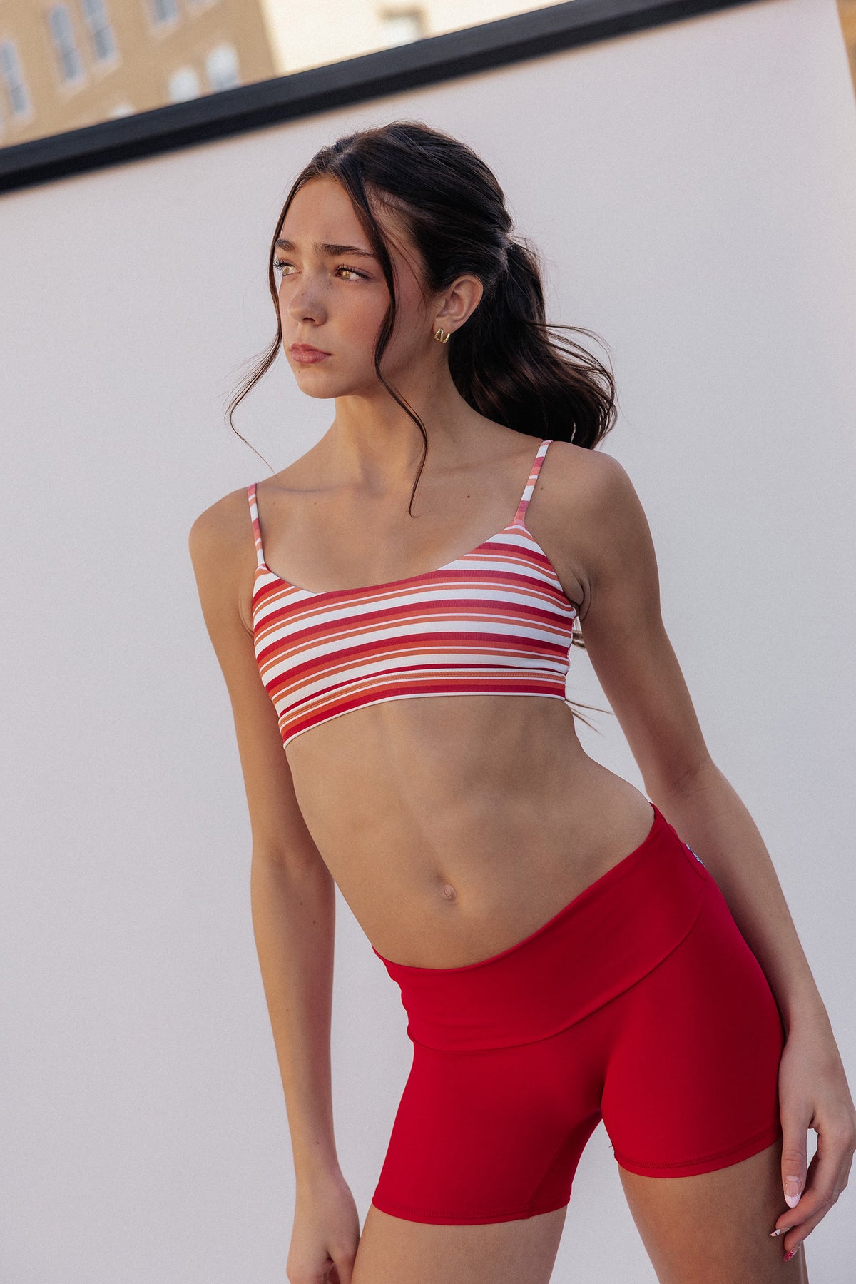kids, modeling a red and white striped sports bra and matching red athletic shorts against a plain white wall.