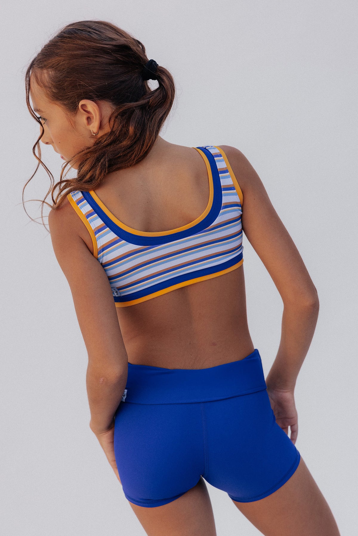 kids,  shown from the back wearing a striped athletic sports bra with blue, white, and yellow trim, paired with royal blue athletic shorts, against a light gray studio background.