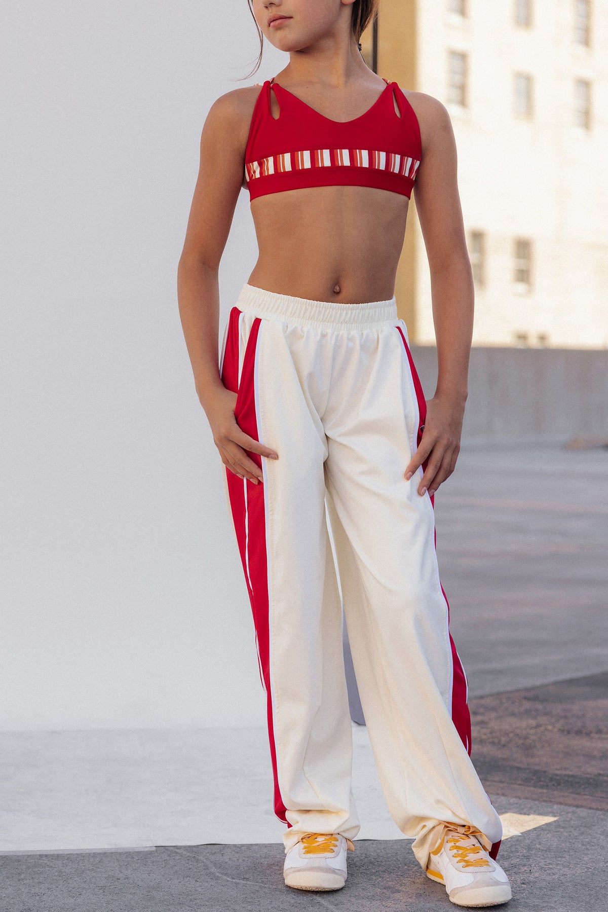 kids, A model wearing a red sports bra with white stripes and white sweatpants with red side stripes, standing outdoors against a light-colored wall.
