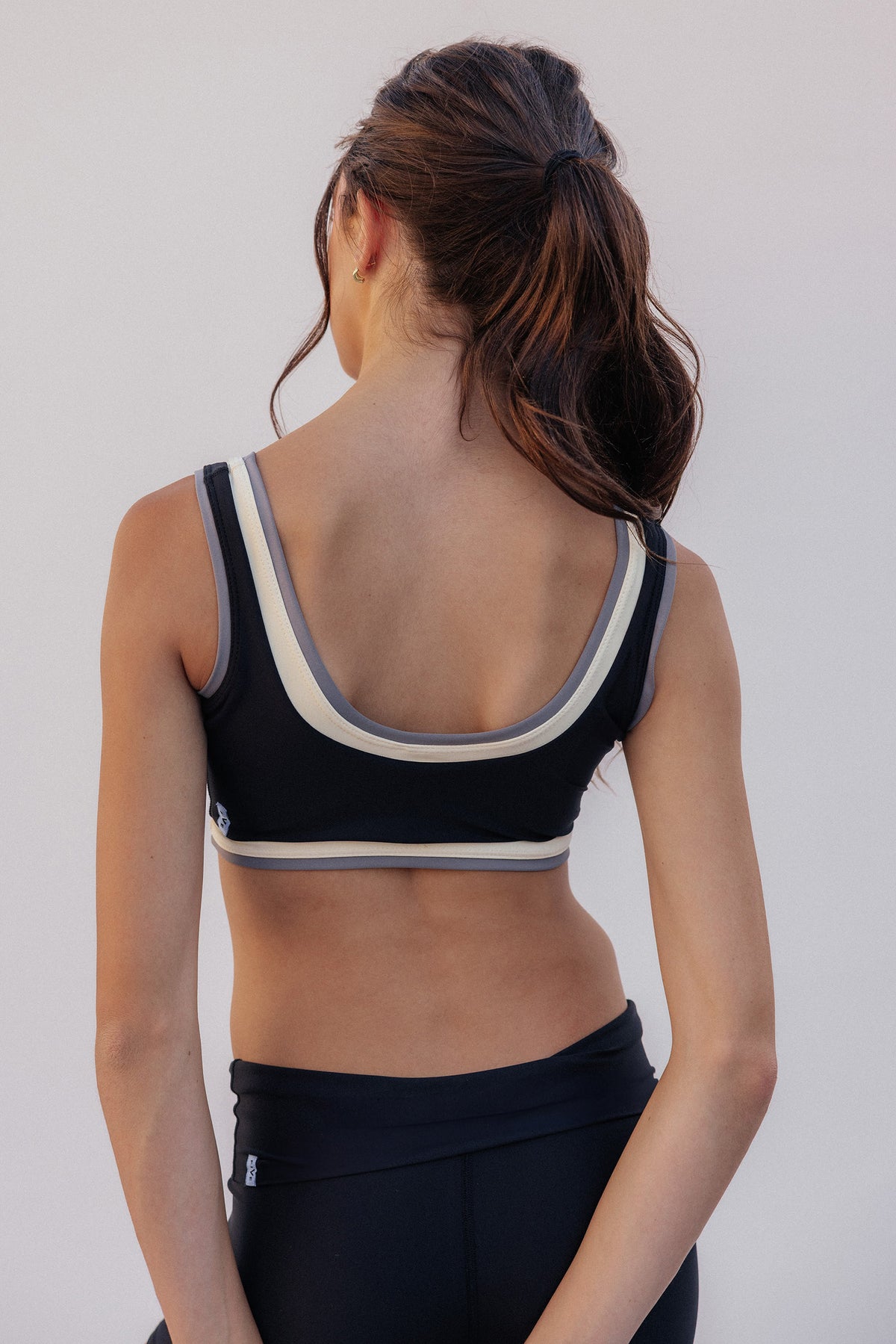 kids, shown from the back wearing a black sports bra with cream and gray trim, paired with black athletic leggings, against a plain light gray background.