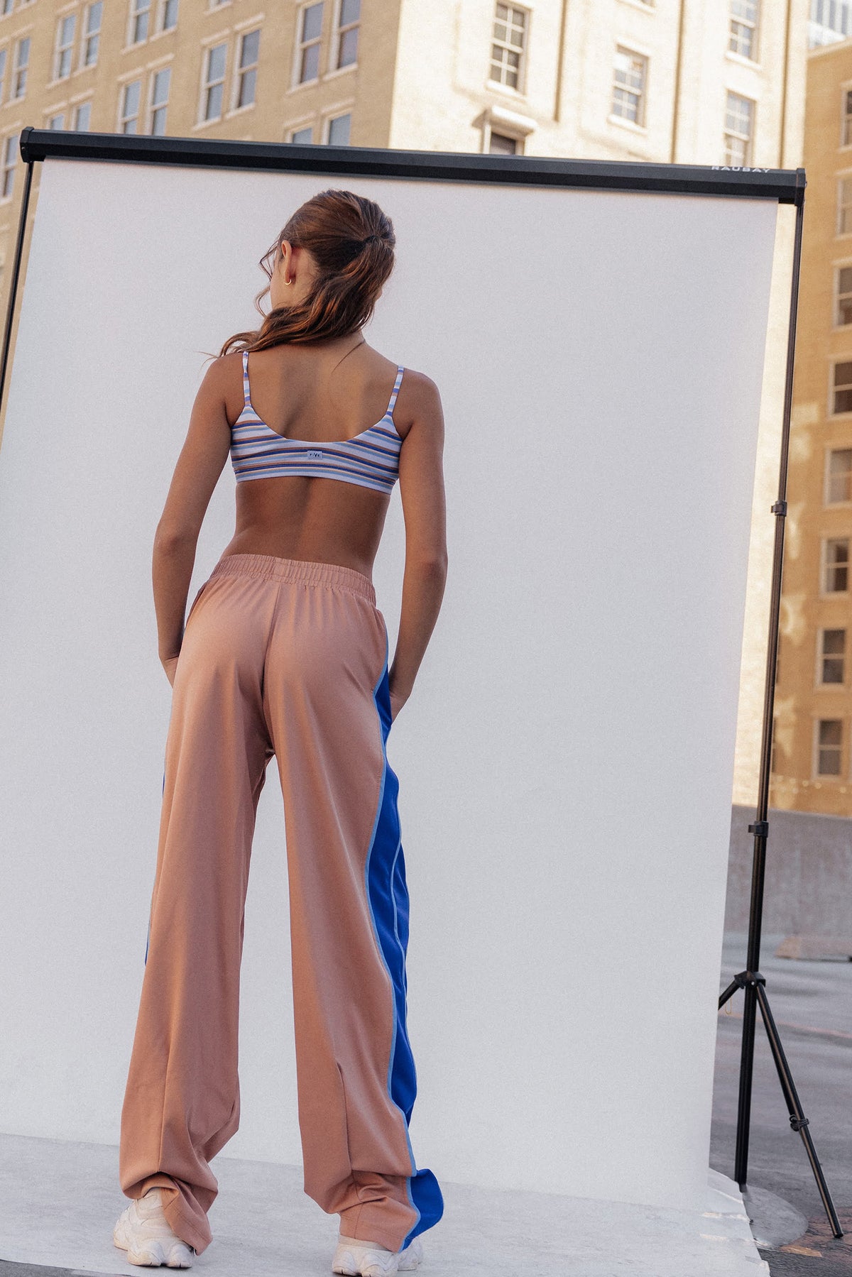 kids, A woman stands with her back to the camera wearing a striped sports bra and loose peach-pink pants with a blue side panel, posed in front of a white studio backdrop with buildings visible in the background.