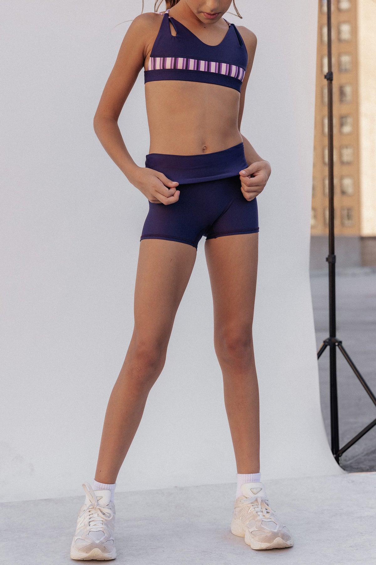 kids, A girl modeling a matching athletic set consisting of a dark purple sports bra with a pink and white striped band and matching dark purple athletic shorts, paired with white sneakers.