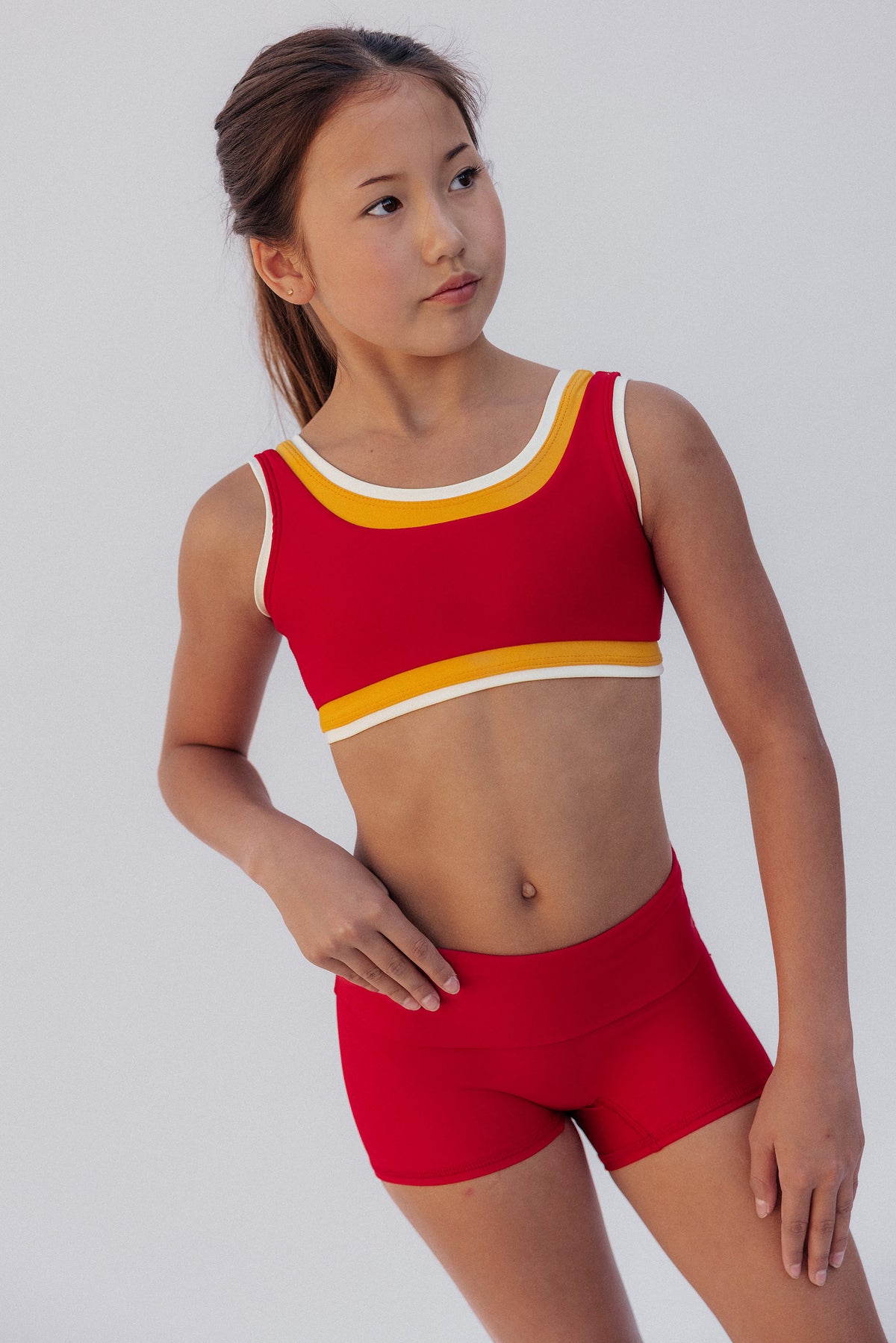 kids, A young girl modeling a red two-piece athletic set consisting of a scoop-neck crop top with yellow and white trim, and matching red athletic shorts, against a light gray studio background