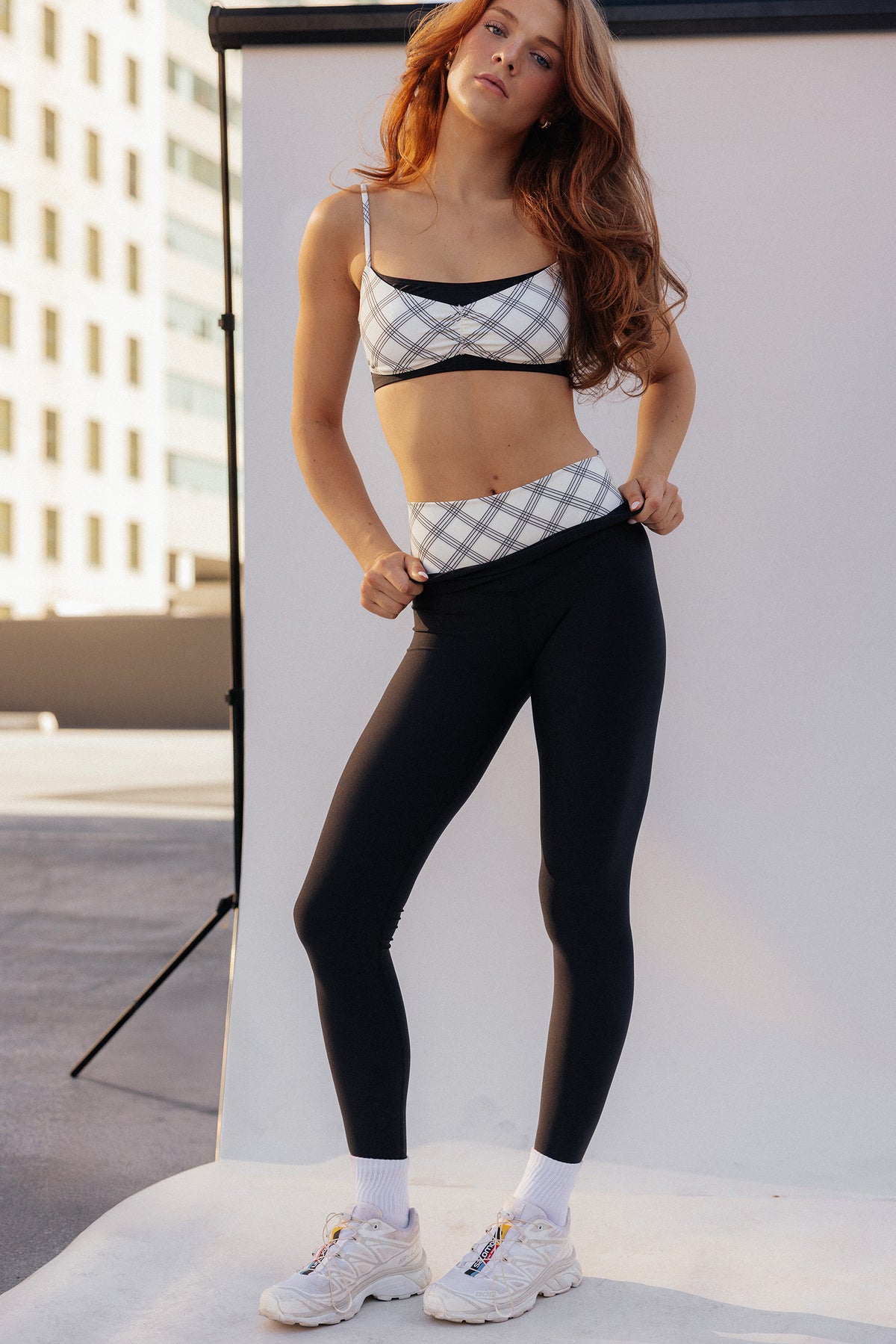 teen,  modeling a matching activewear set consisting of a black and white plaid pattern sports bra and high-waisted black leggings, paired with white socks and white sneakers, set against a white studio backdrop.