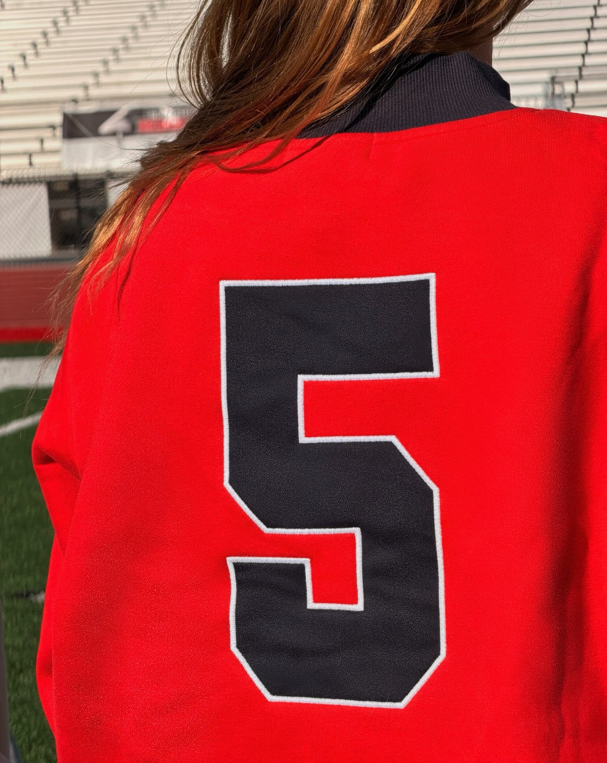 kids, A close-up back view of a girl wearing a bright red varsity-style jacket with a large navy number 5 outlined in white on the back, photographed outdoors near stadium seating and a field.