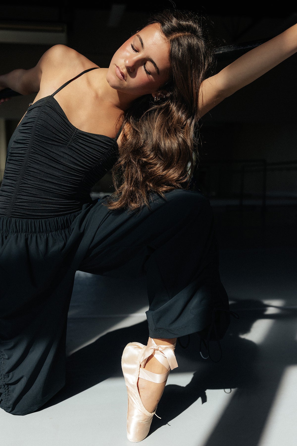 teen, A girl in a black dance Leo poses dynamically on pointe in a ballet slipper, with strong sunlight casting shadows on a dark studio floor.