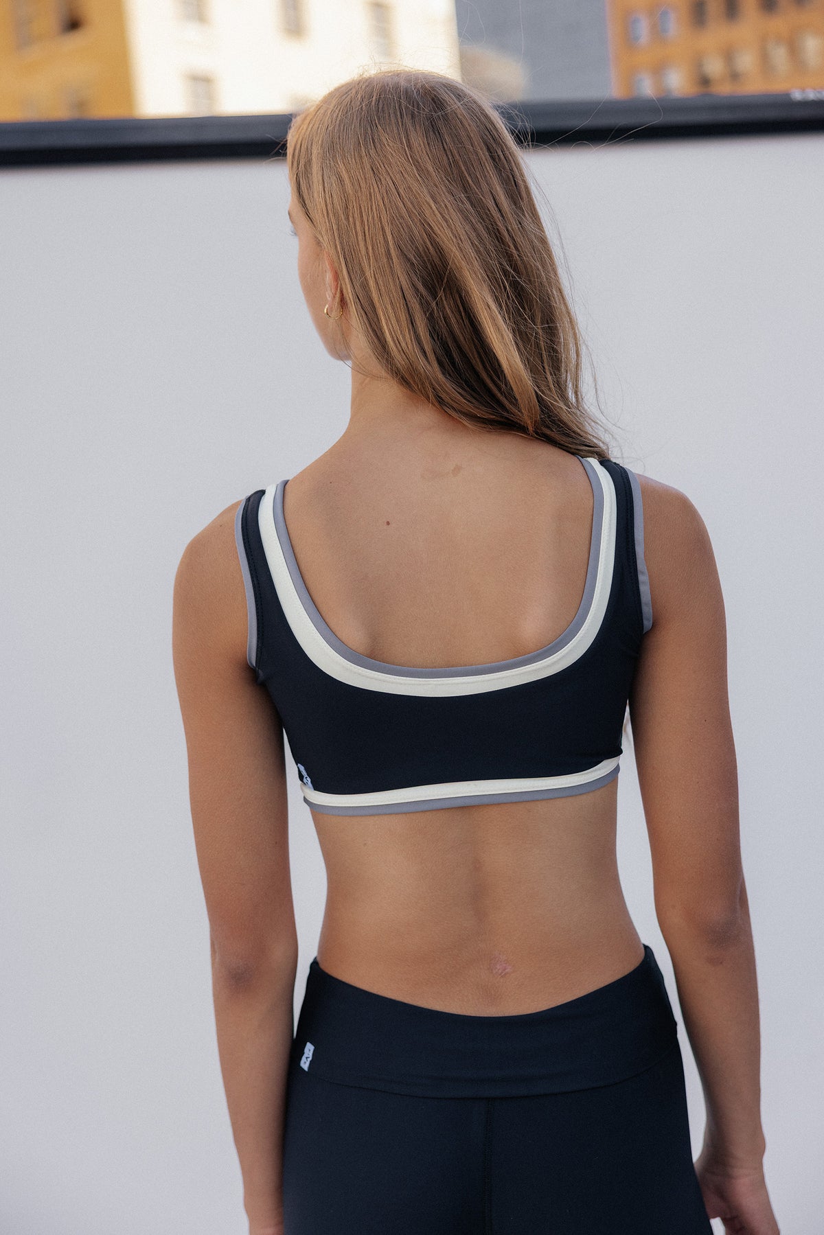 teen, viewed from behind wearing a black sports bra with white and gray trim and black leggings, standing against a white wall with buildings in the background.