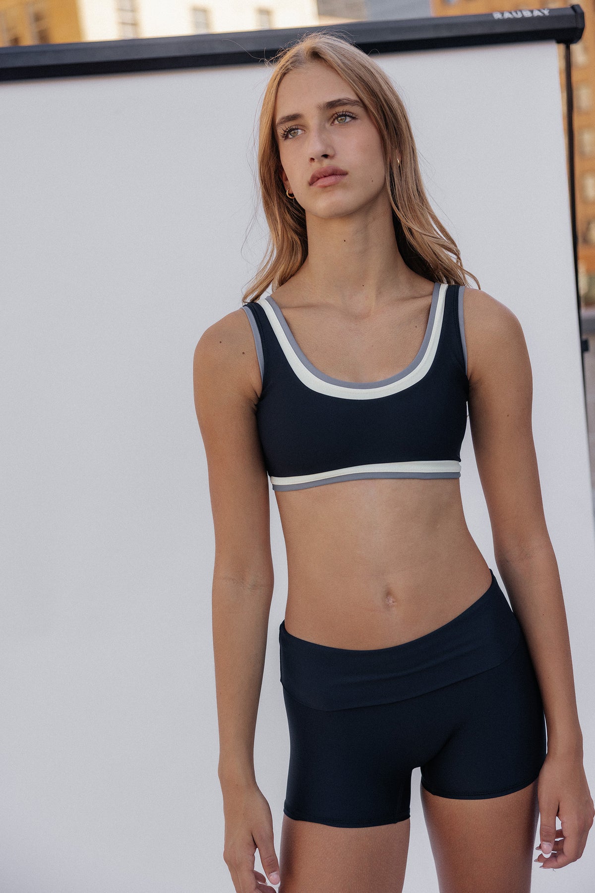 teen, modeling a matching athletic set consisting of a scoop-neck sports bra and high-waisted shorts in black with white and gray trim, standing against a white studio backdrop.
