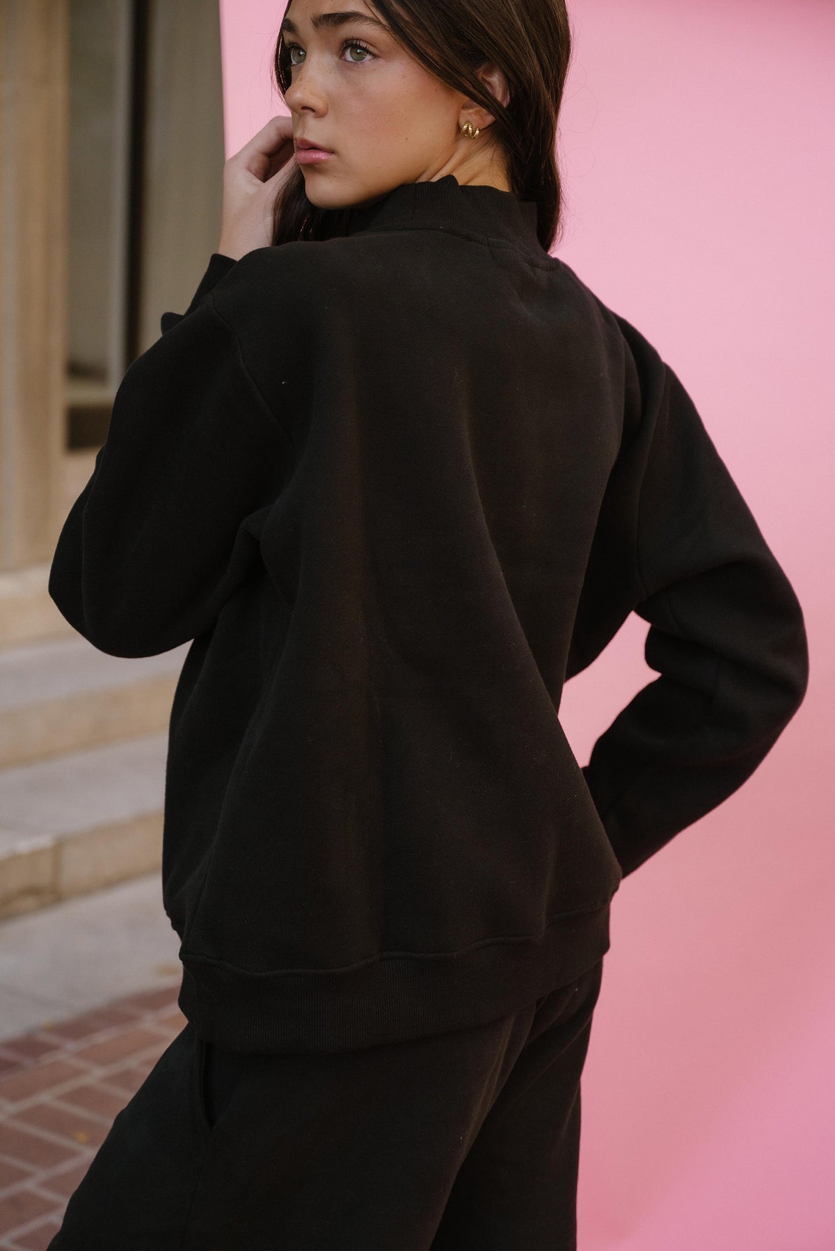 kids, a girl wearing a black crewneck sweatshirt and matching sweatpants, photographed from the back against a vibrant pink wall.