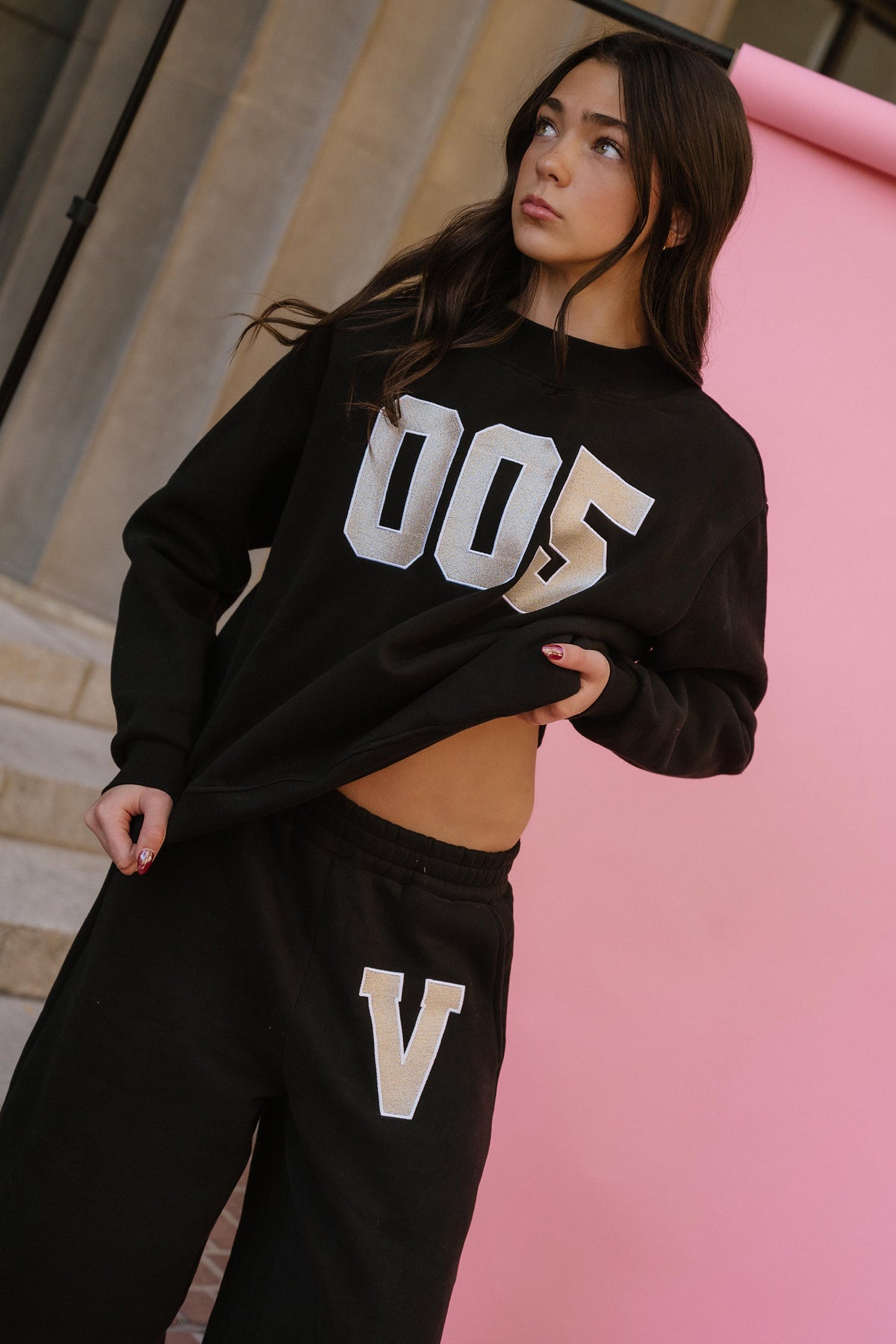 kids, A girl wearing a black sweatshirt with large silver varsity-style '005' lettering on the front and matching black sweatpants with a large white 'V' on the thigh, posed against a pink wall.