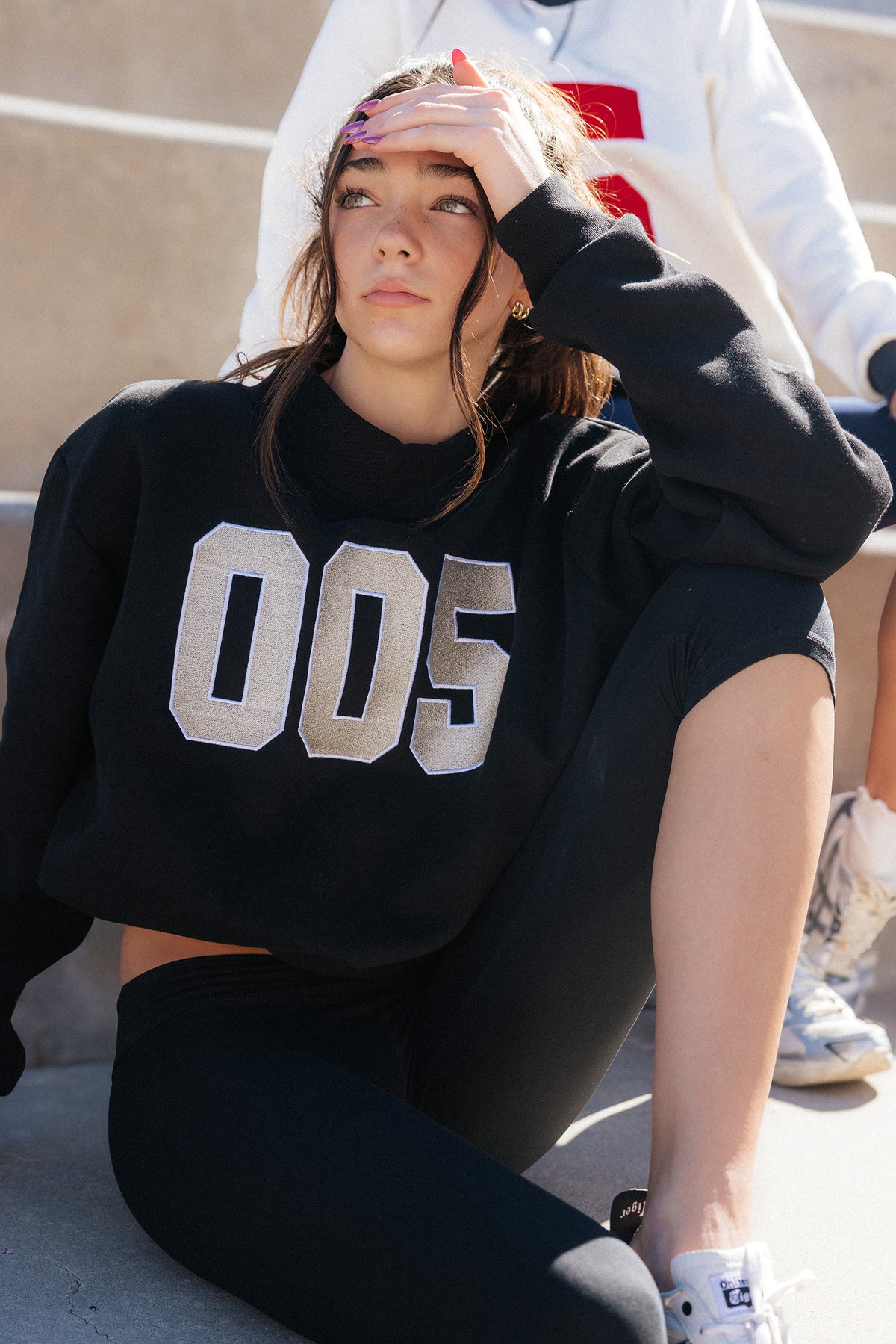 kids, A girl sitting outdoors wearing a black cropped sweatshirt with large metallic silver '005' lettering on the front, paired with black leggings and white sneakers.