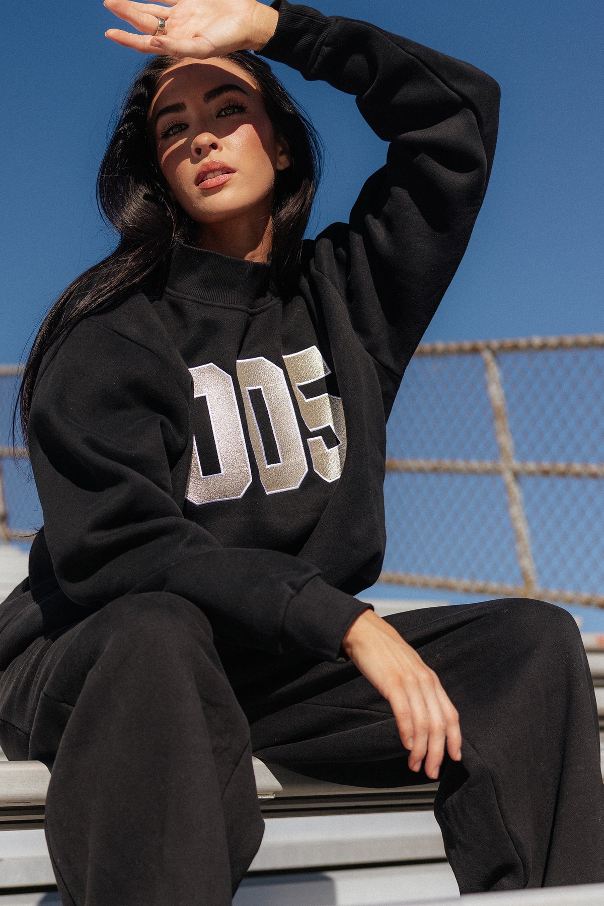teen, A girl wearing a black sweatshirt and matching sweatpants sits on outdoor bleachers under a blue sky. The sweatshirt features a large metallic silver '005' graphic on the front.