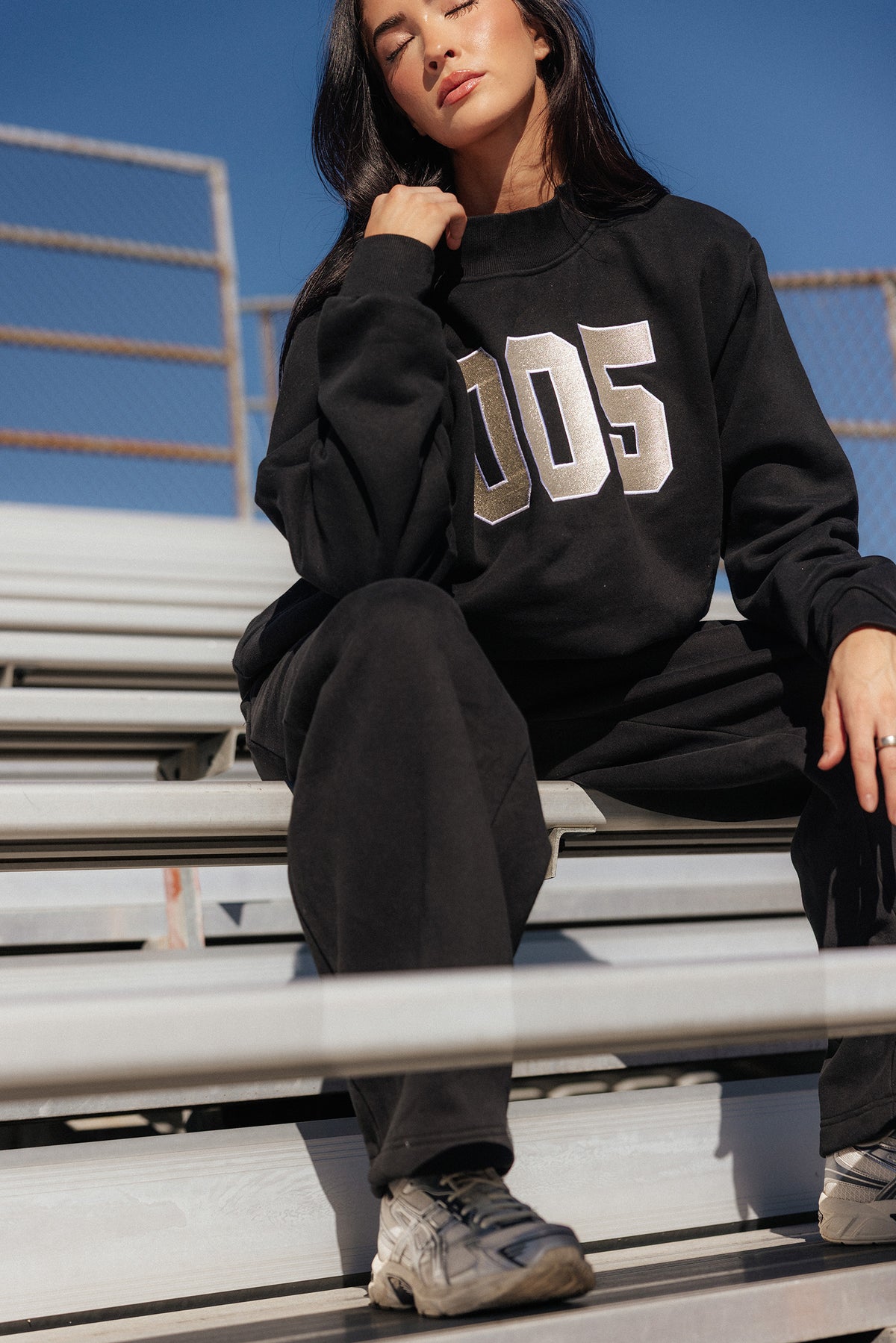 teen, A girl sitting on outdoor metal bleachers wearing a black sweatshirt with large metallic silver '005' lettering on the front, paired with black sweatpants and gray sneakers.