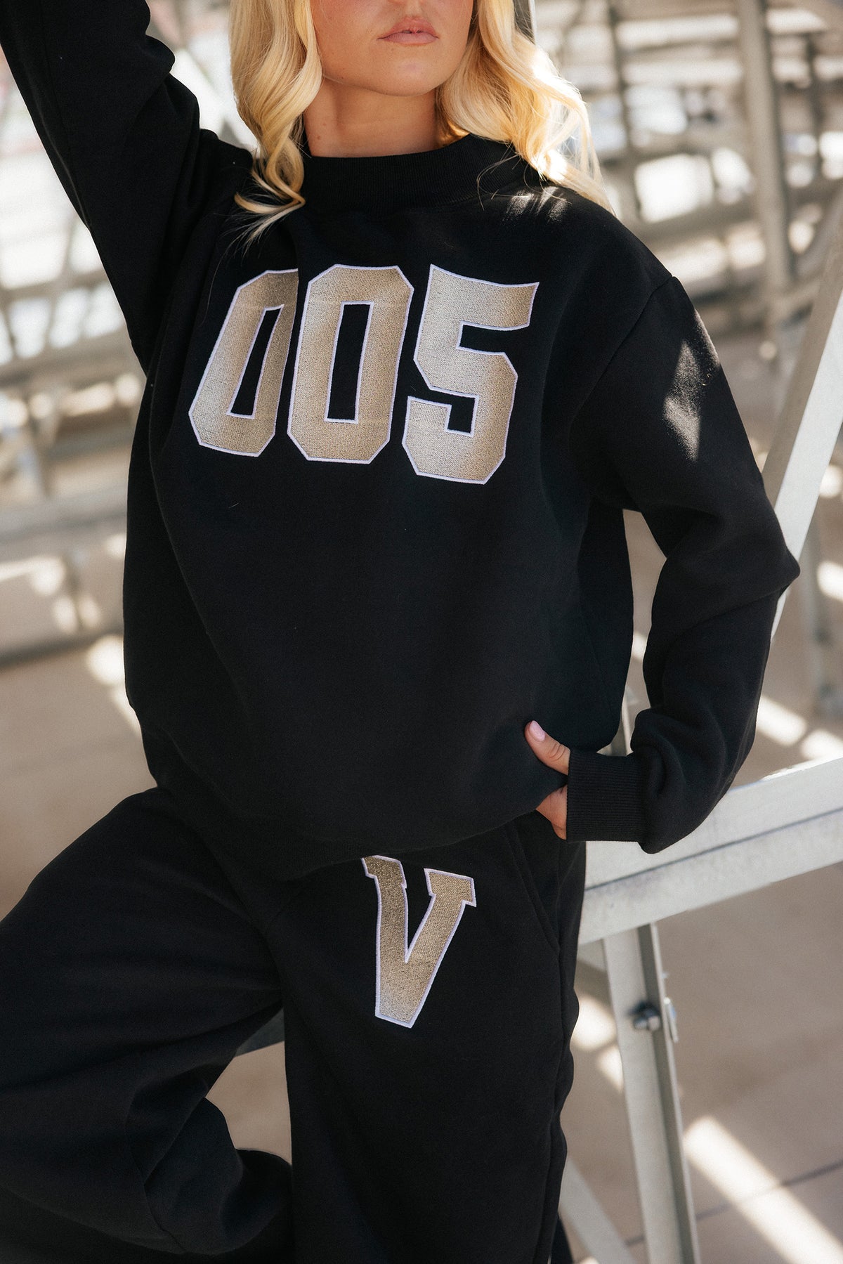 teen, A girl wearing a black sweatshirt and sweatpants set with large beige and silver varsity-style lettering, photographed in an outdoor setting with stadium seating in the background.