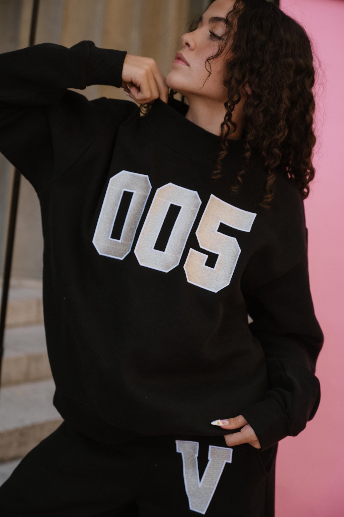 teen, A girl wearing a black sweatshirt with large metallic silver block numbers '005' on the front, posed against a pink wall and stone background.